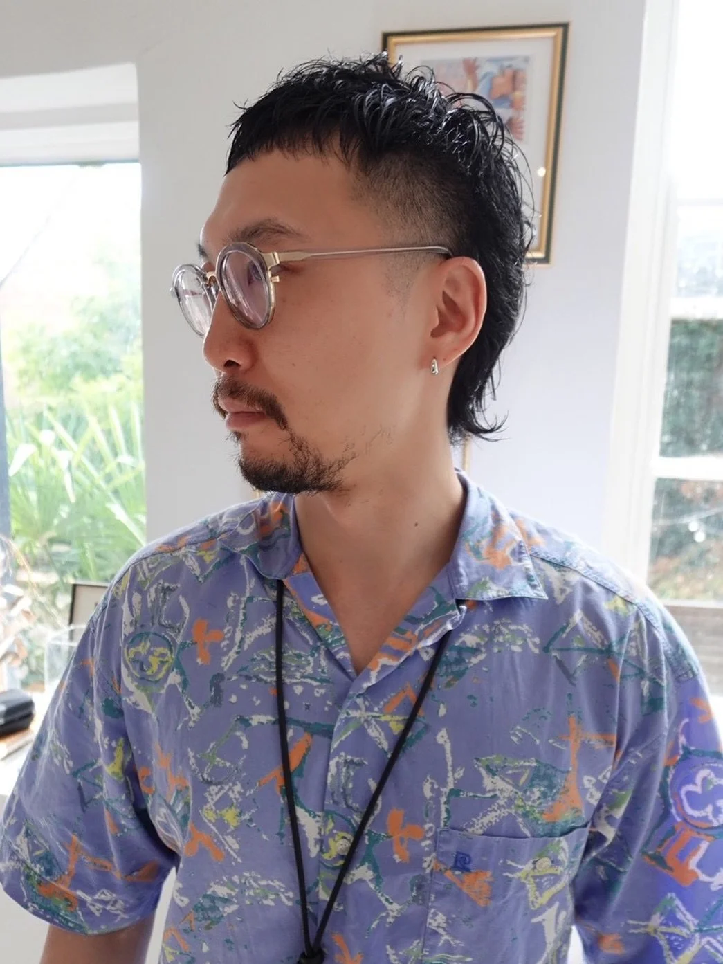 Profile of a man with curly black hair, sunglasses, a colorful patterned shirt, and an earring, indoors with windows and a framed picture in the background.