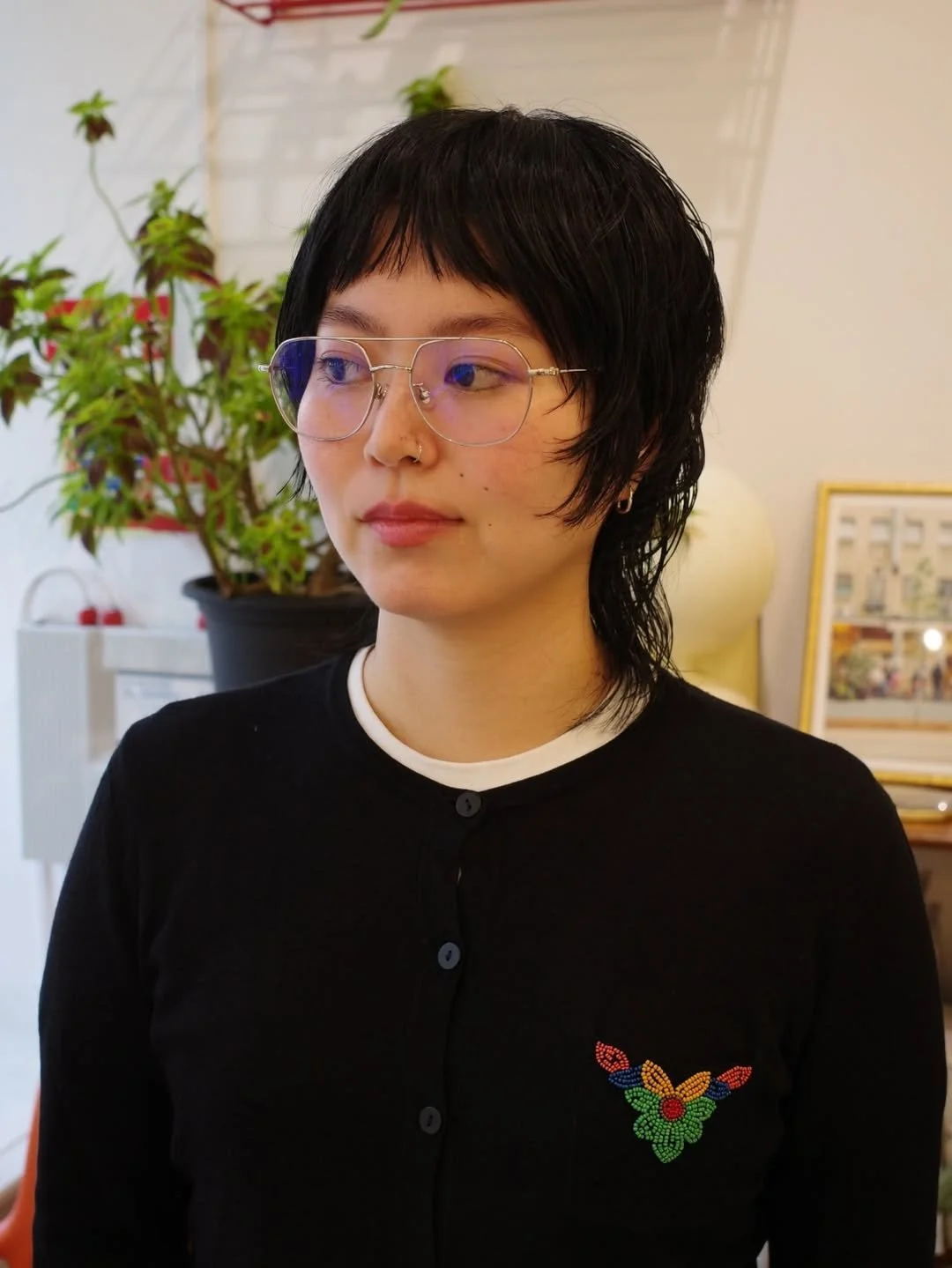 A woman with short black hair, glasses, and a nose ring. She is wearing a black cardigan with a colorful beaded butterfly design on the chest. She is in an indoor space with plants and framed pictures in the background.