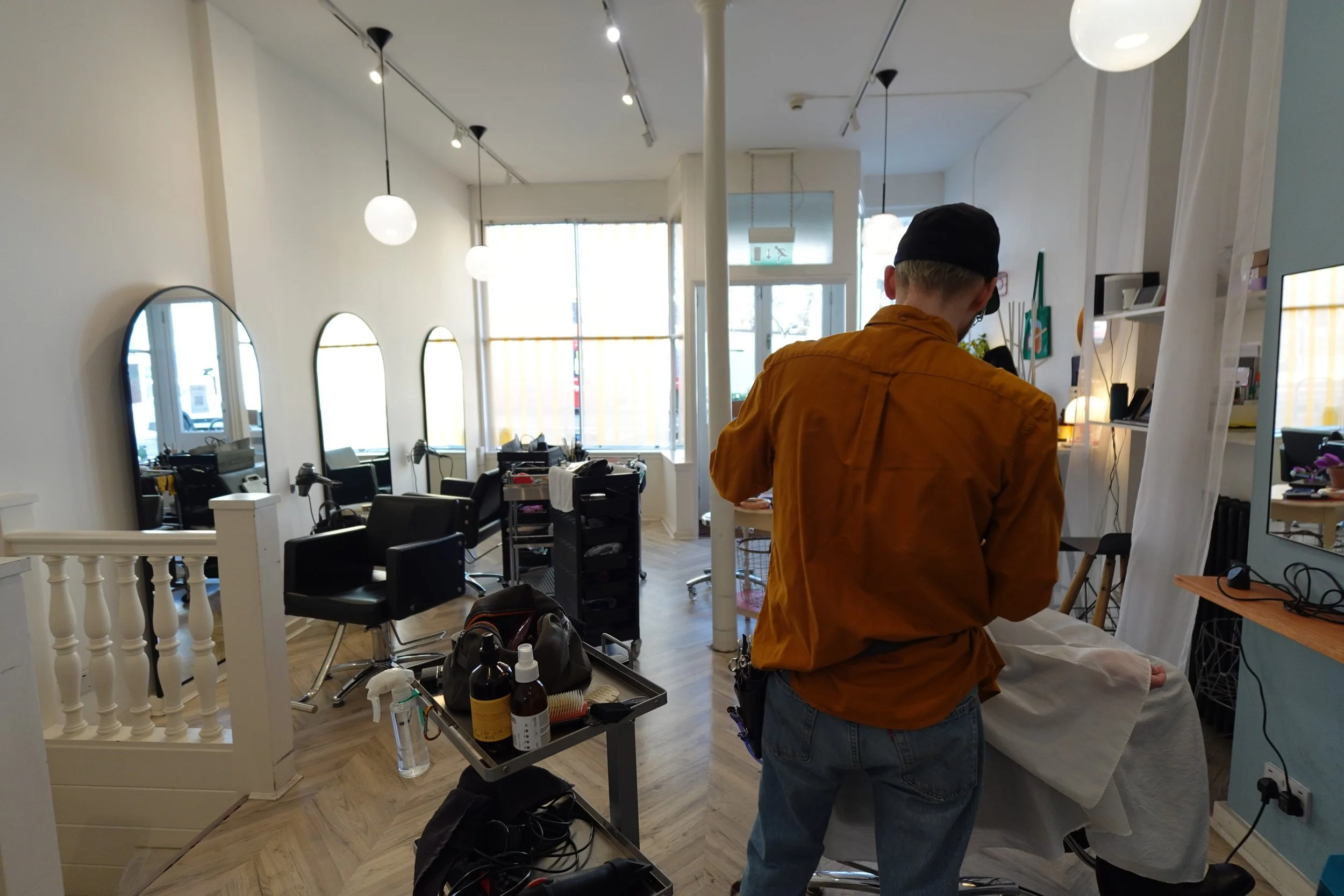 A person in an orange shirt and jeans giving a haircut to a customer in a salon. The salon is well-lit with multiple mirrors, chairs, and hairdressing tools visible.