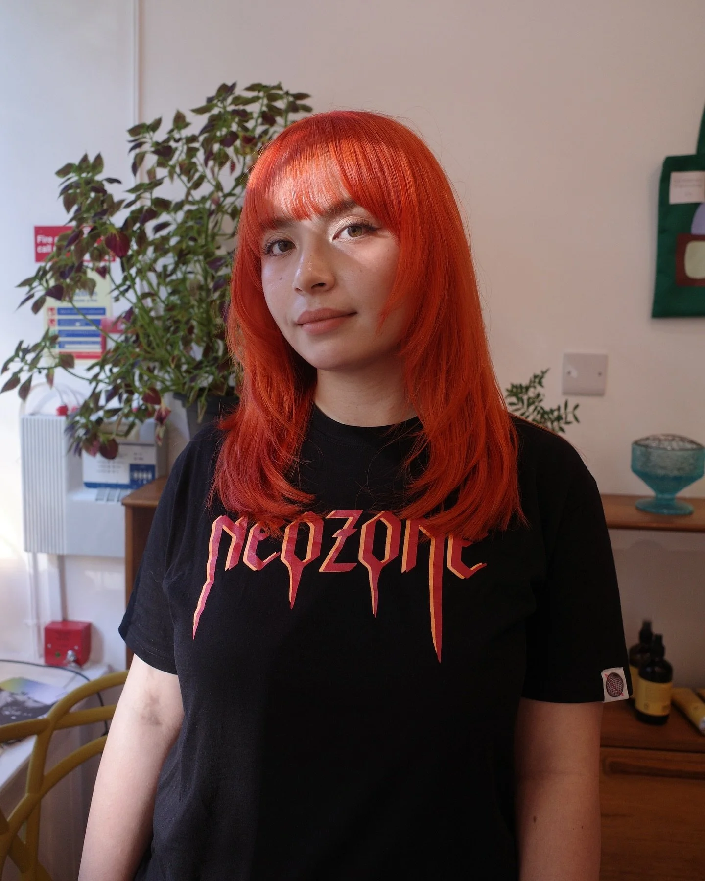 A young woman with bright red hair and fair skin, wearing a black T-shirt with a colorful graphic, standing indoors in front of a white wall with a plant and household items in the background.