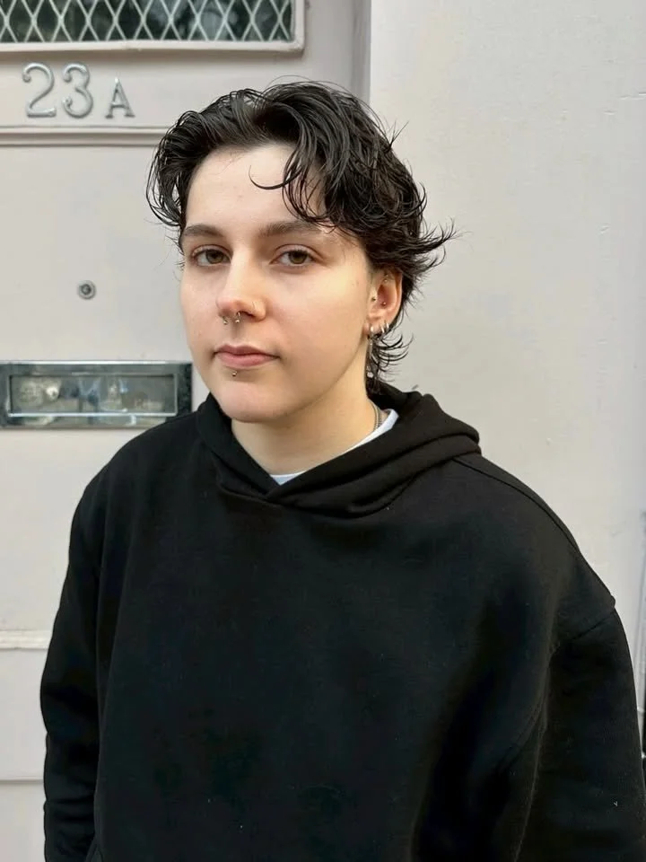 A young person with short, wavy dark hair, light skin, and facial piercings wearing a black hoodie, standing outside in front of a beige wall with the address number 23A.