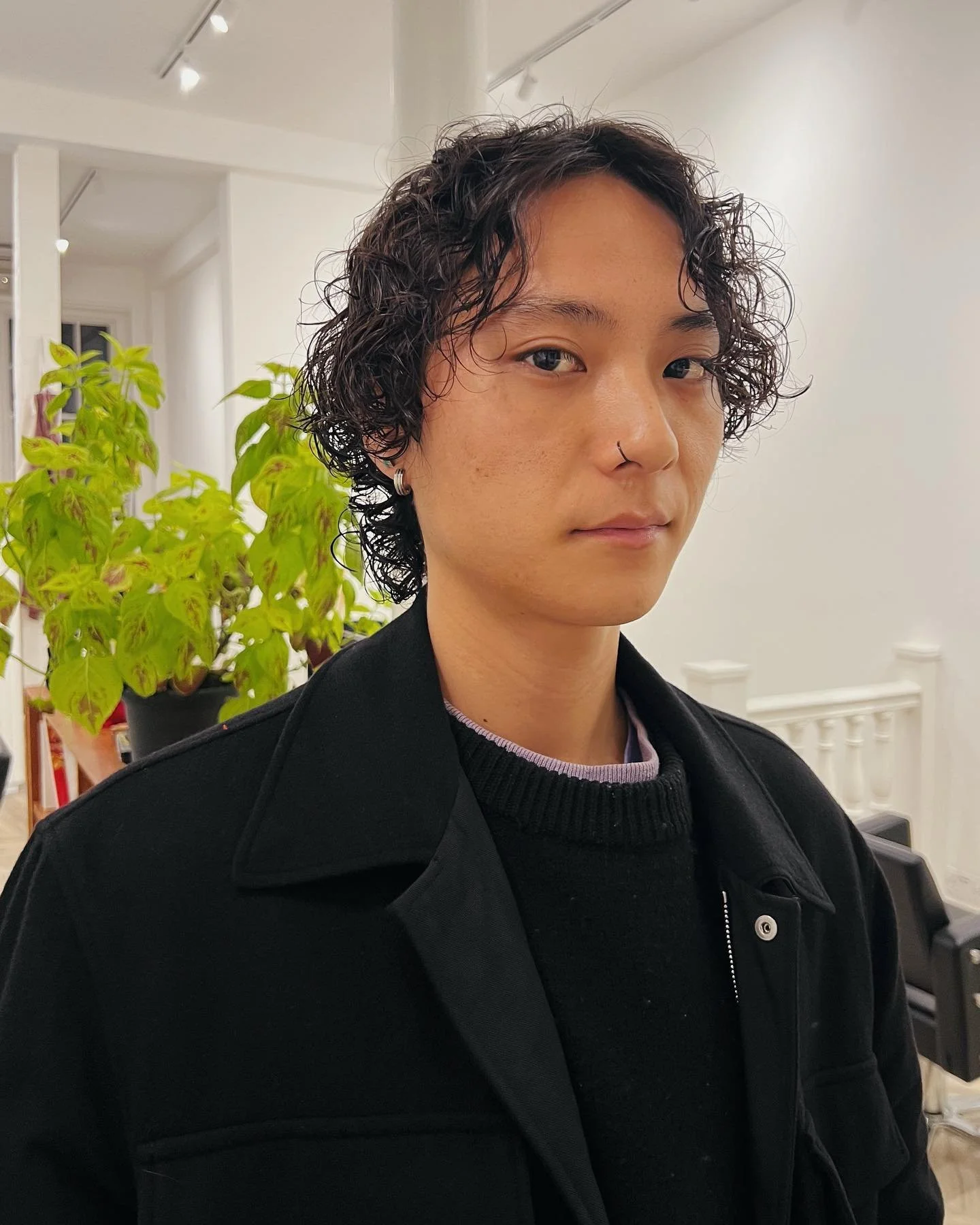 Portrait of a person with short curly black hair, wearing a black jacket and silver earrings, standing indoors near a green potted plant.