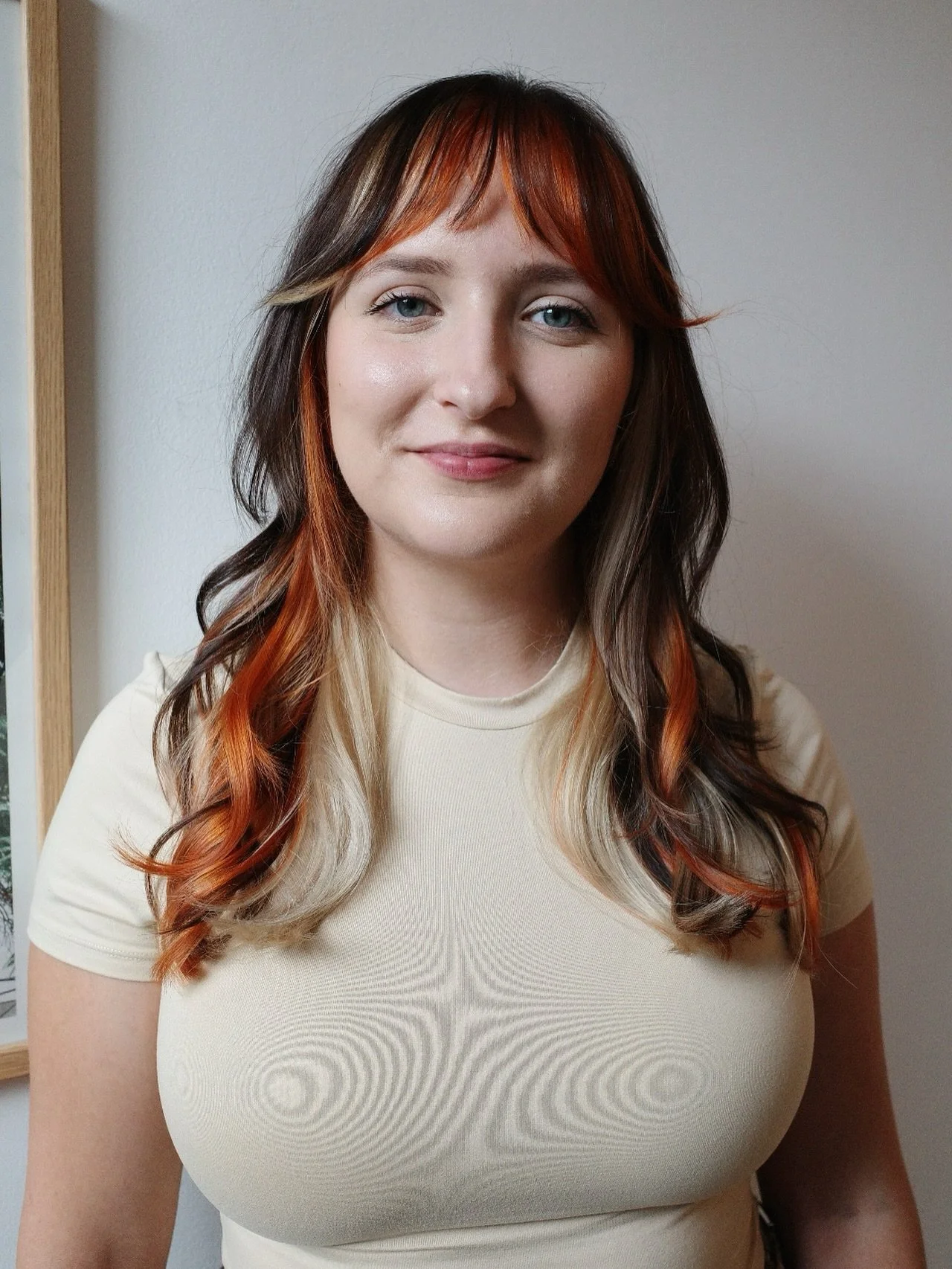 A woman with shoulder-length hair with red and blonde highlights, wearing a beige t-shirt, standing in front of a white wall with a window to her left.
