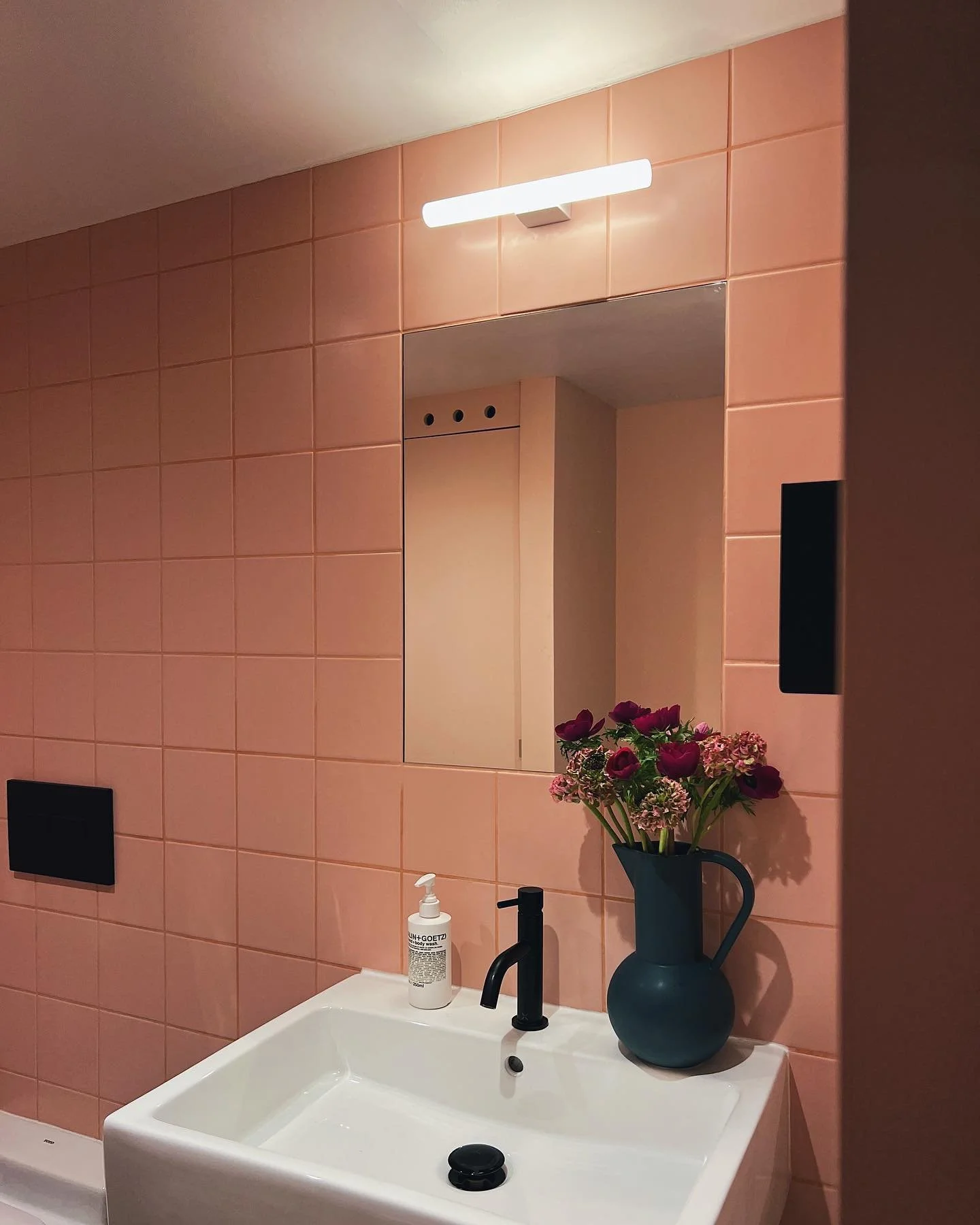 Bathroom with pink tiled wall, white sink, black faucet, soap dispenser, and a dark vase with pink and purple flowers.