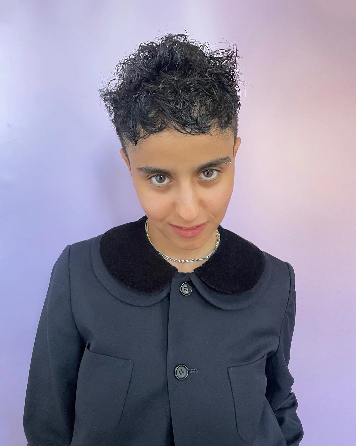 Young person with short, curly dark hair and light skin, wearing a black jacket with a rounded collar, standing against a lavender wall.