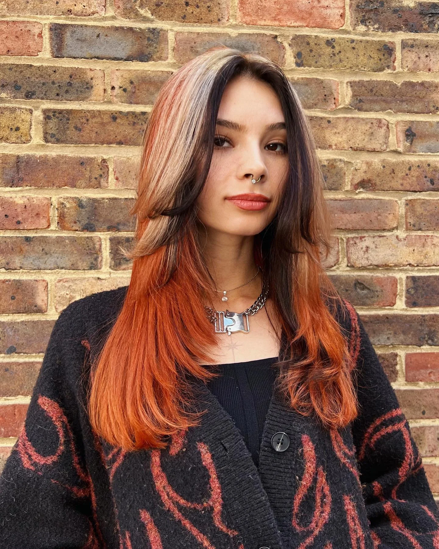 A young woman with long, orange and brown hair standing against a brick wall. She is wearing a black cardigan with red designs, a black top, necklaces, and has a septum piercing.