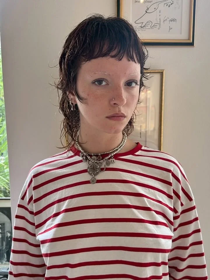 Young person with short dark hair, wearing a red and white striped shirt, layered necklaces, and earrings, standing indoors in front of a framed picture on the wall.