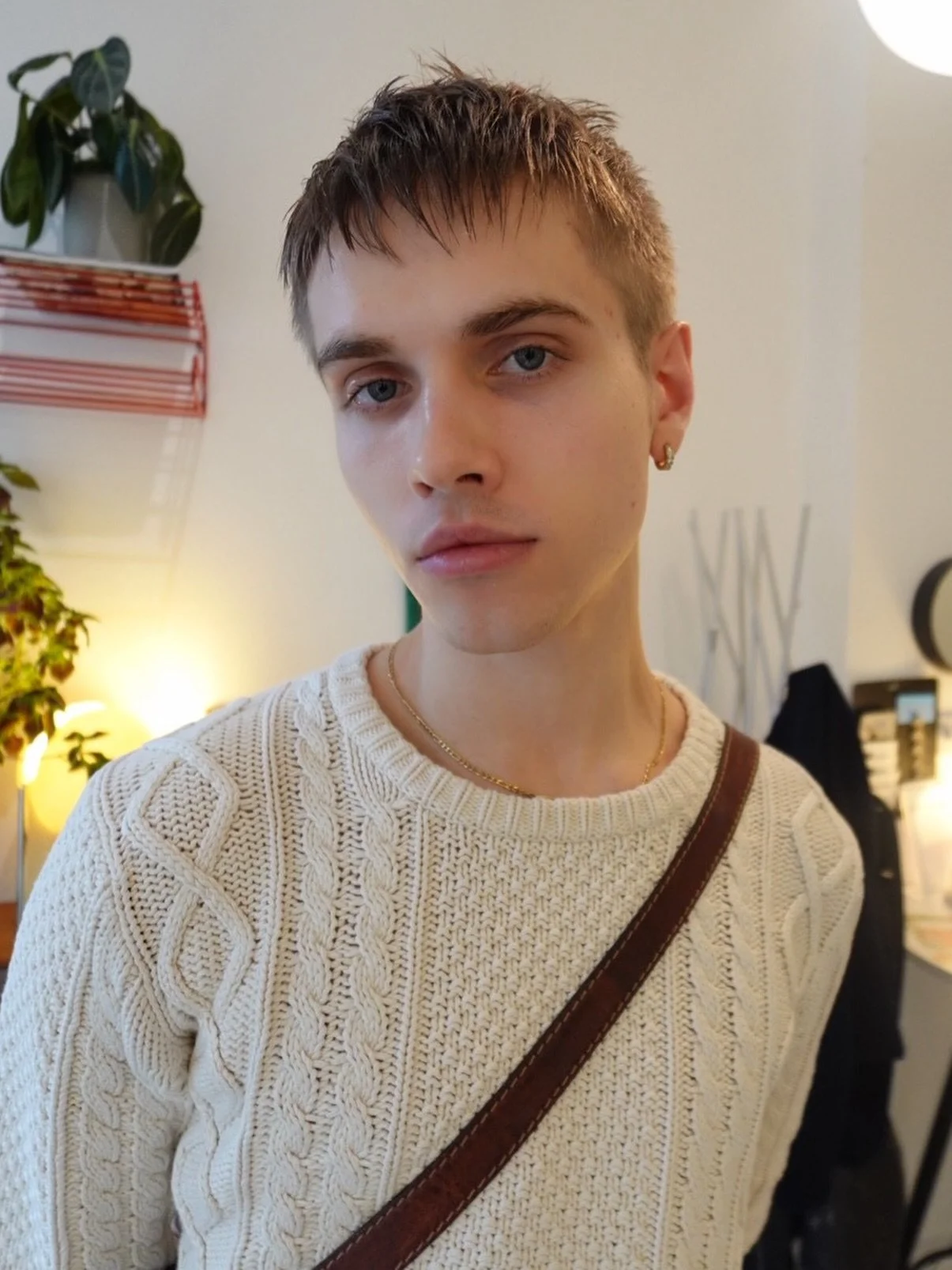 A young man with light skin, short brown hair, and blue eyes taking a selfie indoors. Wearing a cream-colored knit sweater, gold hoop earrings, and a gold chain, with a brown crossbody strap visible.