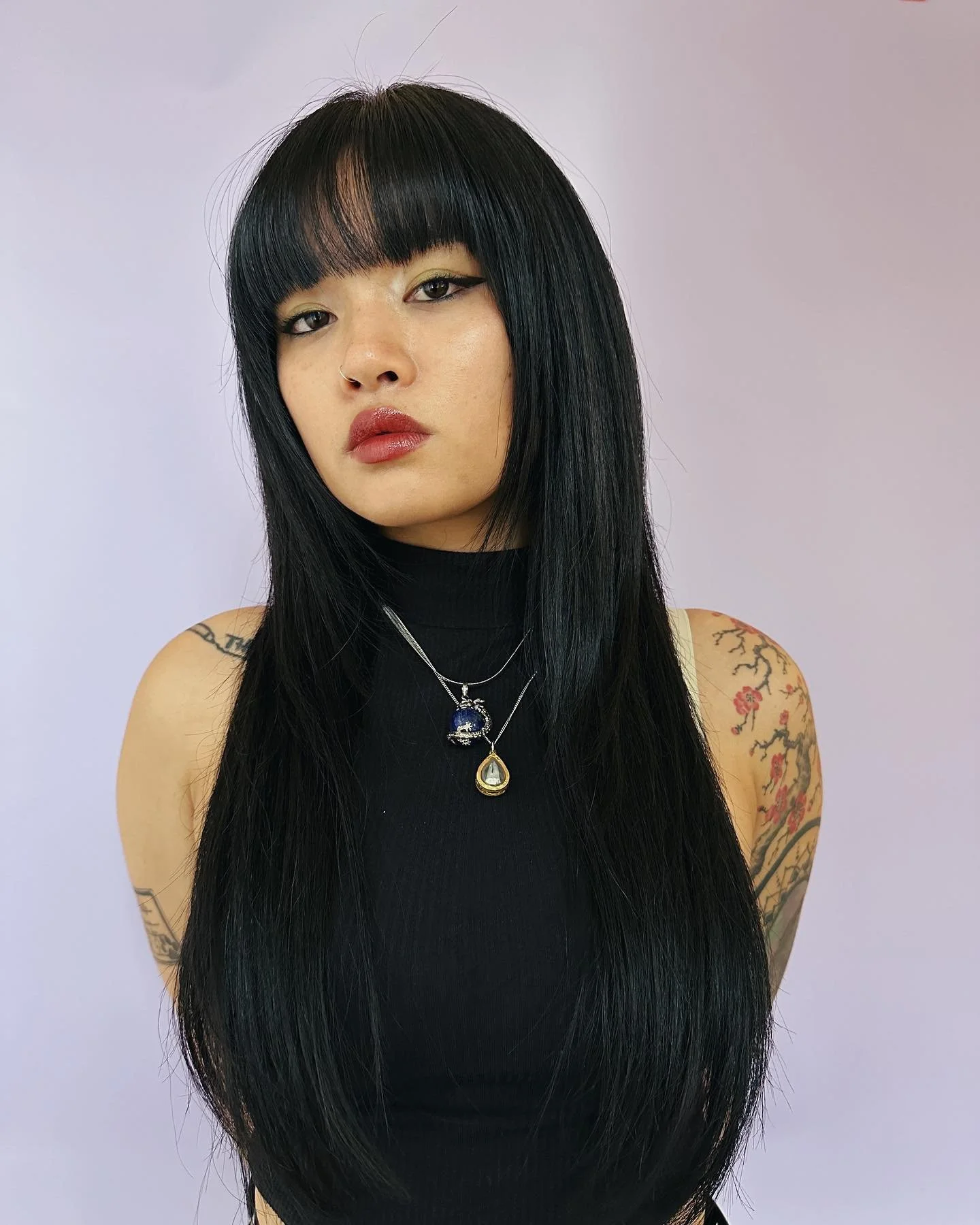 A woman with long black hair, wearing a black sleeveless top, and layered necklaces, looking directly at the camera against a plain light-colored background.