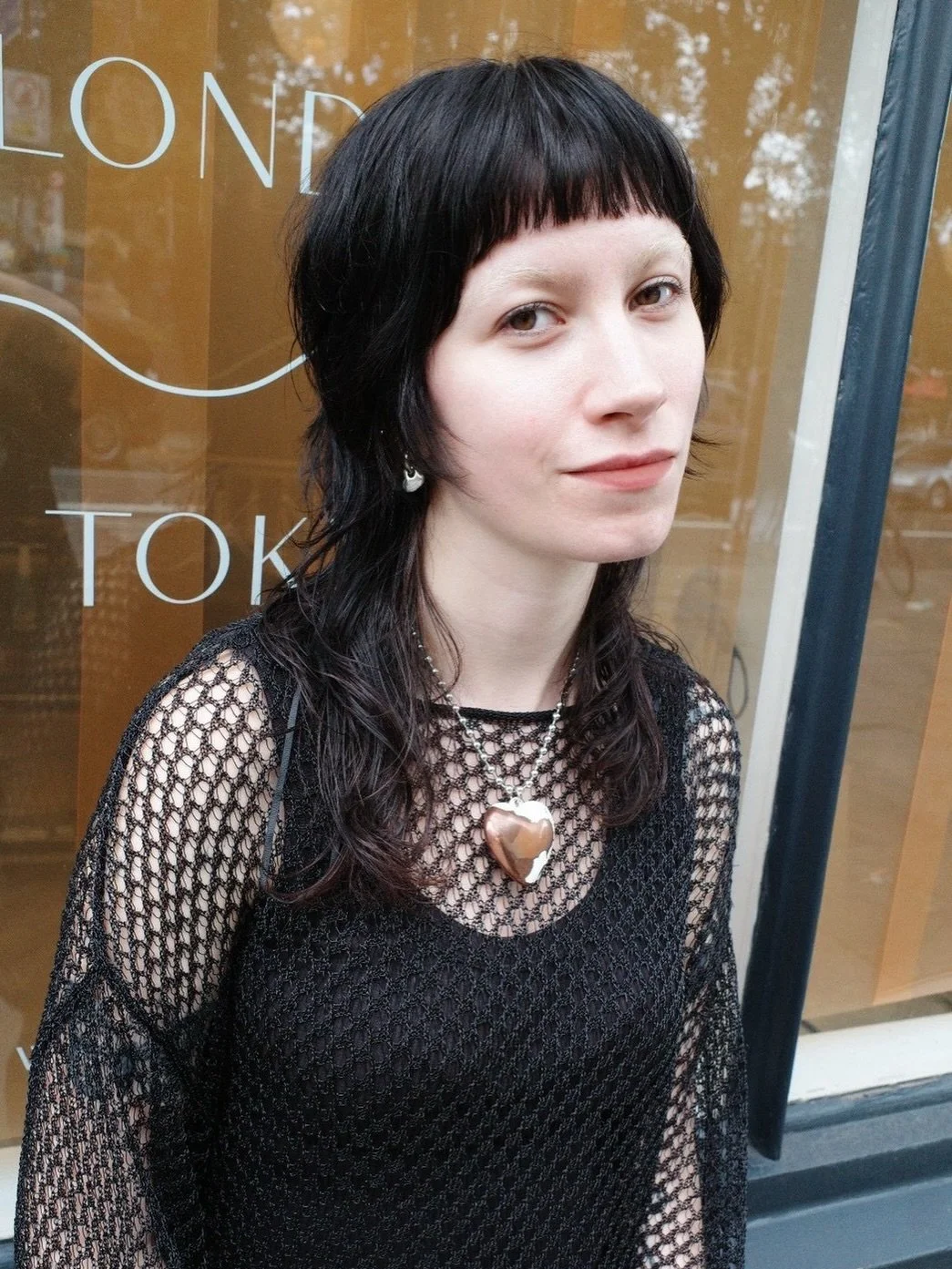 Young woman with black hair and bangs wearing a black fishnet top and a large heart-shaped pendant necklace, standing in front of a glass window with white lettering.