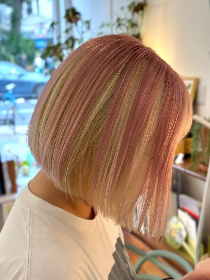 Close-up of a person with a short, straight bob haircut dyed in pastel pink and blonde shades, indoors with a blurred background of plants and furniture.