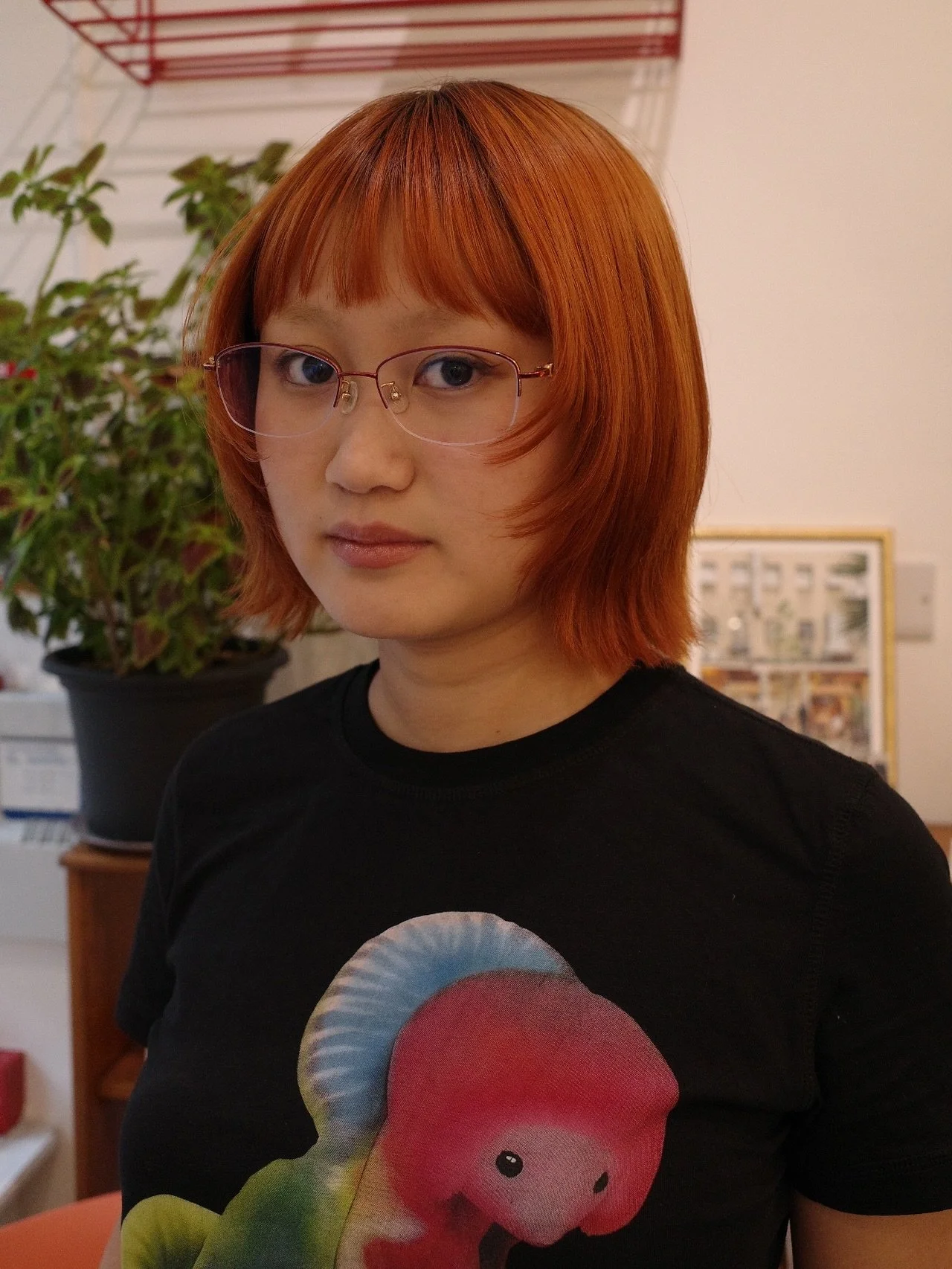 A woman with short red hair and glasses standing indoors, wearing a black T-shirt with a colorful cartoon dinosaur on it, behind her a large potted plant and framed pictures on the wall.