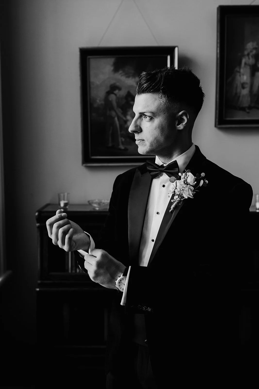 Black and white photograph of a man in a tuxedo adjusting a cufflink, featuring a boutonnière, standing in front of wall art.