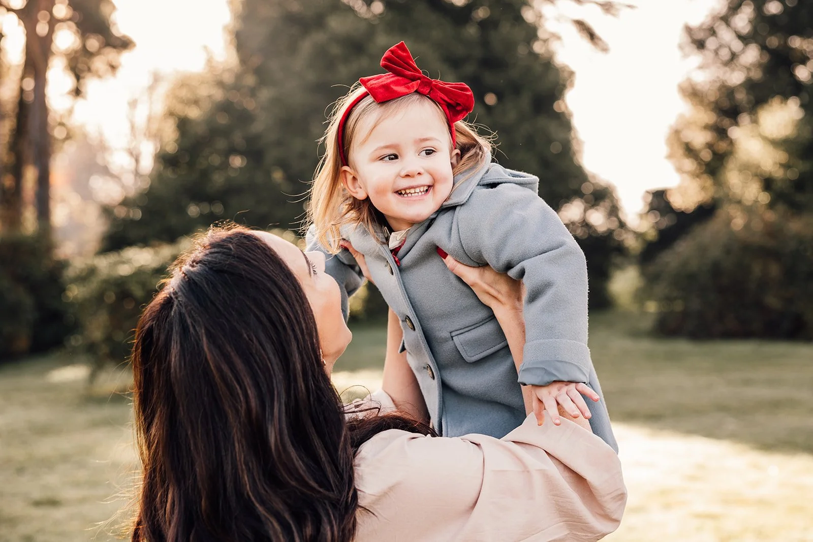 Mother lifting smiling toddler with red bow outdoors, surrounded by trees and greenery.