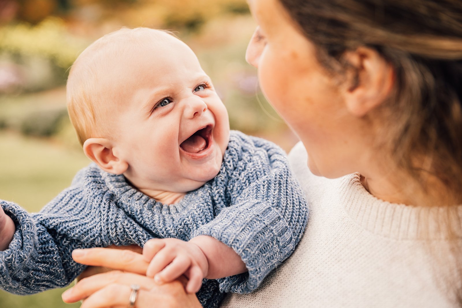 A smiling baby in a blue sweater being held by a woman outdoors.