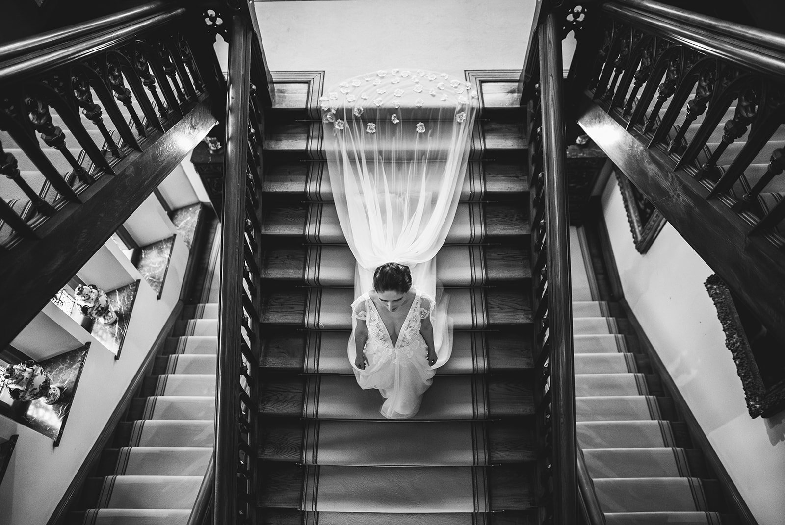 A bride in a wedding dress ascending a staircase with dark wooden railings, viewed from above, with a sheer train and floral details.