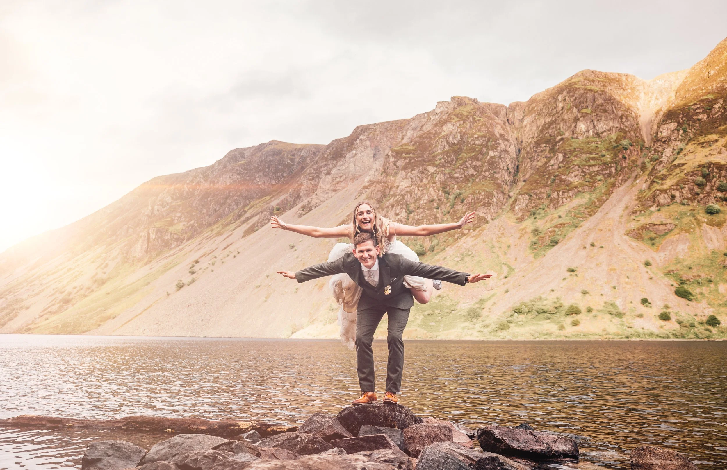 A couple in wedding attire stands on rocks by a lake, with mountains in the background. The woman is on the man's back with arms outstretched, both smiling.