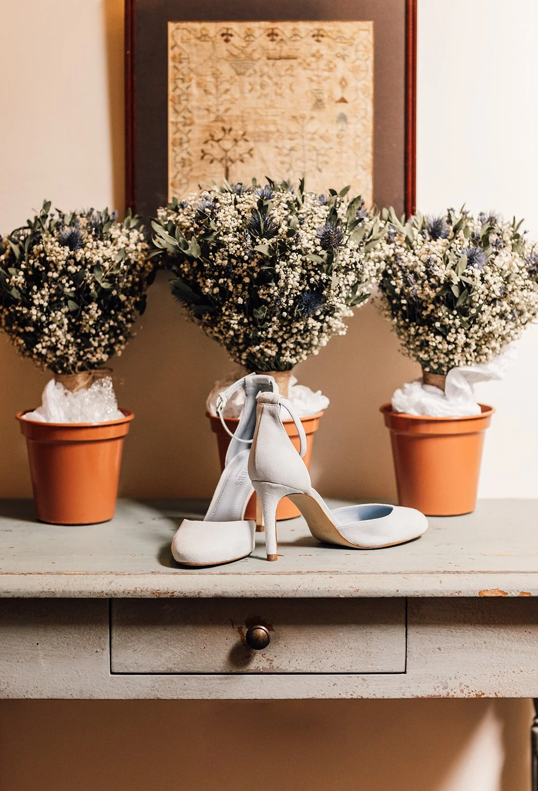 A pair of white high-heeled shoes on a rustic table with three potted flower arrangements in the background.