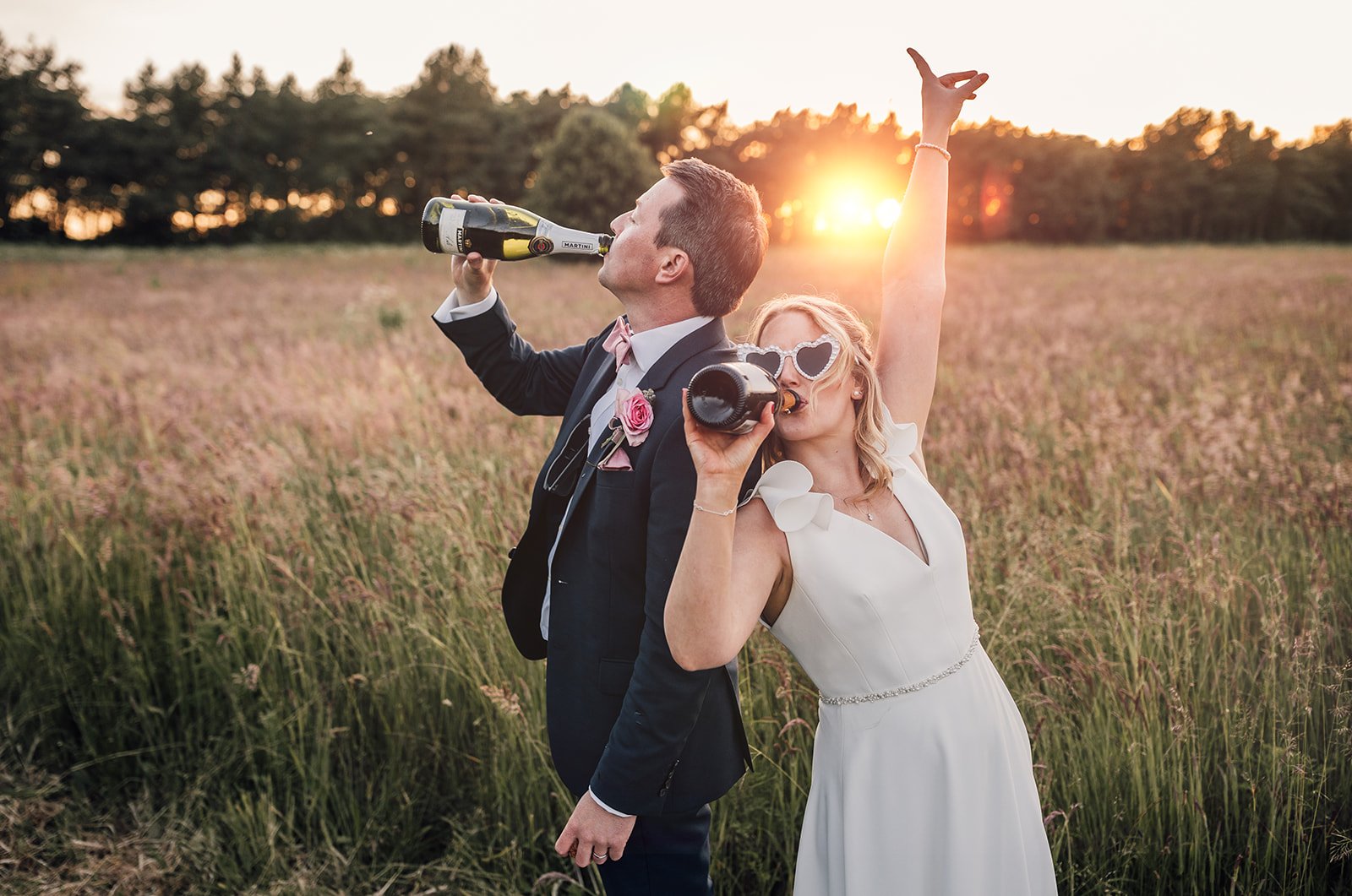 A couple in wedding attire in a field, drinking from champagne bottles at sunset. The bride is wearing a white dress and heart-shaped sunglasses, and the groom is in a suit with a tie and boutonniere.