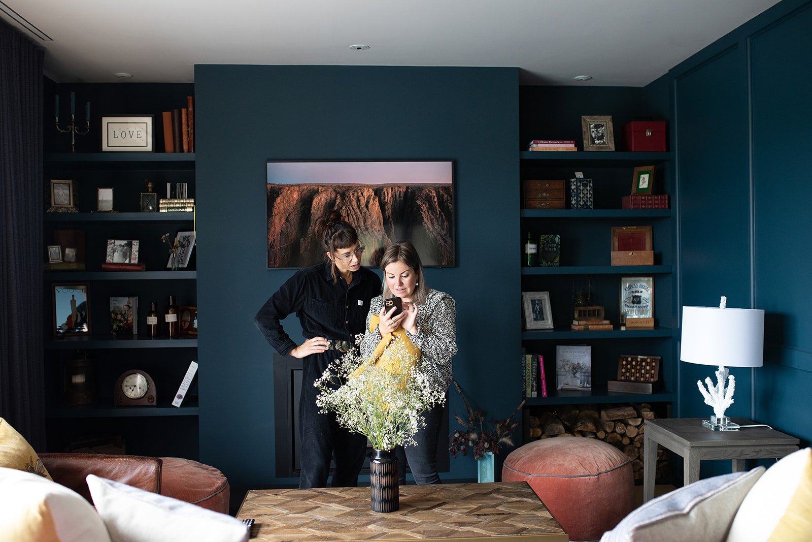 Two women standing in a living room, looking at a phone together. The room has dark blue walls, built-in bookshelves with books and decorative items, a framed photo of a waterfall, and a lamp on a side table. A vase with white flowers on a wooden tab