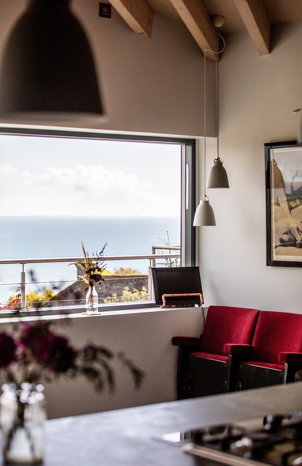 Interior of a room with large window overlooking the ocean, red velvet chairs, framed artwork on the wall, and hanging pendant lights.