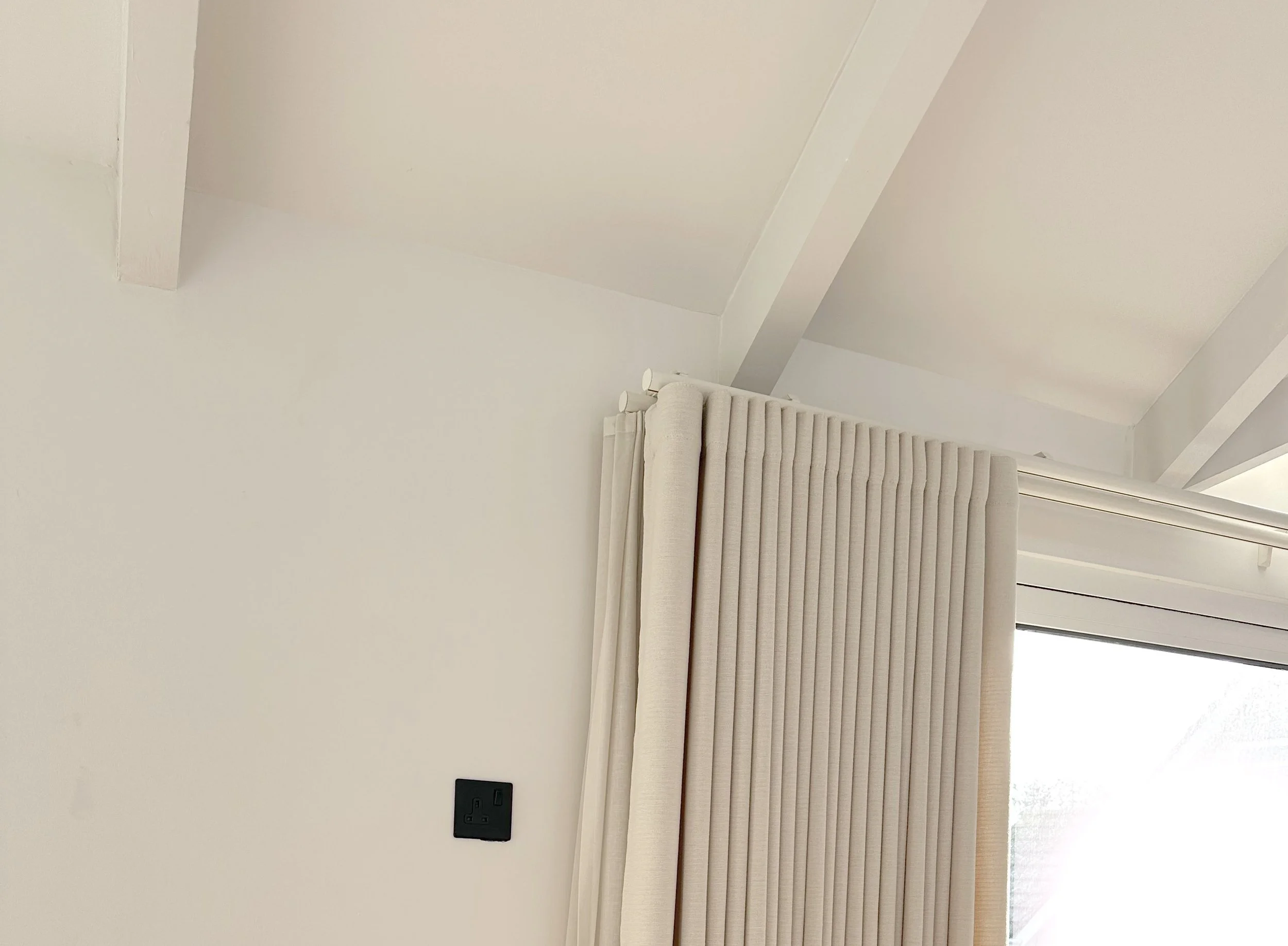 A corner of a room with white walls, beige curtains, and a black electrical socket on the wall.
