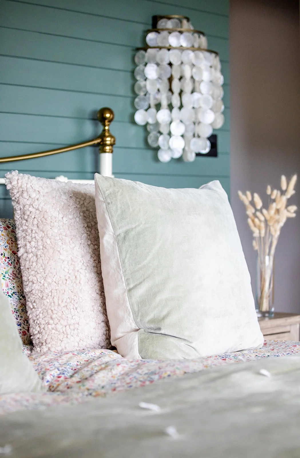 Close-up of a bed with soft pillows, colorful bedspread, a blue wooden wall, and wall decor including a white shell chandelier and a dried flower arrangement in a glass vase.