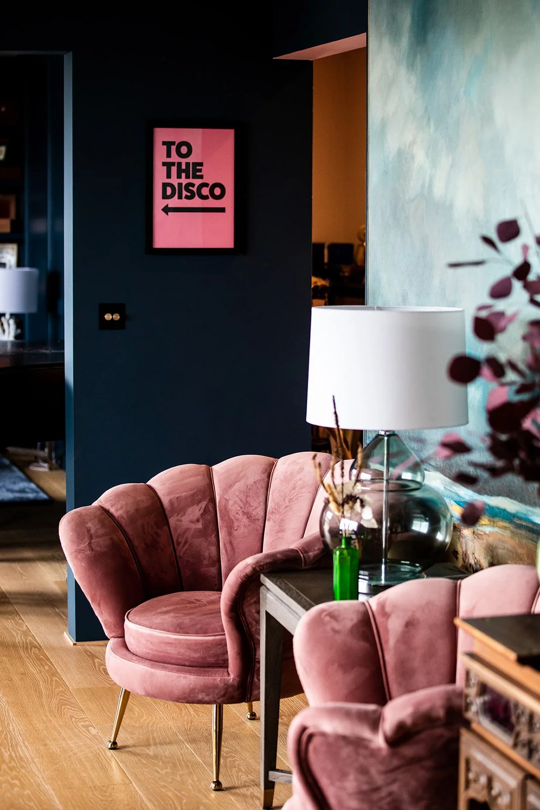 Interior of a stylish room with pink velvet chairs, a table with a lamp, and a sign on the wall that says "TO THE DISCO" with an arrow pointing left.