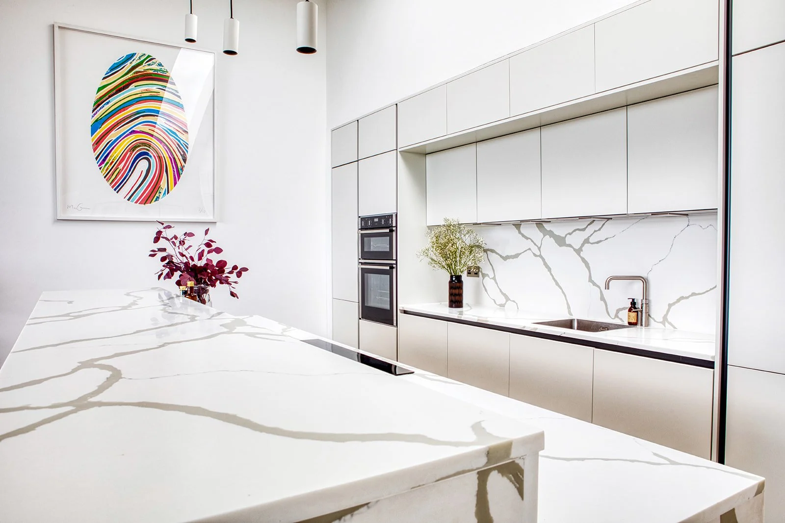 Modern kitchen with white cabinetry, marble countertop, and backsplash, featuring built-in ovens, a sink with a brown soap dispenser, a vase with greenery, and colorful abstract artwork on the wall.