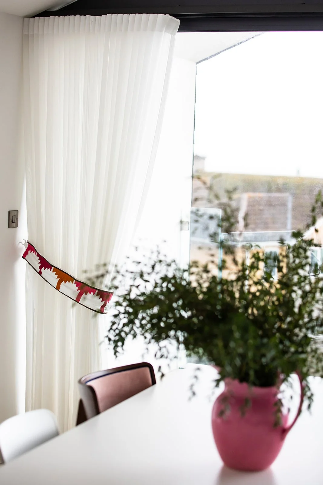 A dining area with a large pink flowerpot holding a green, leafy plant, a white table, and brown chairs. A sheer white curtain partially covers a large window or sliding glass door, with a decorative banner hanging on the curtain.
