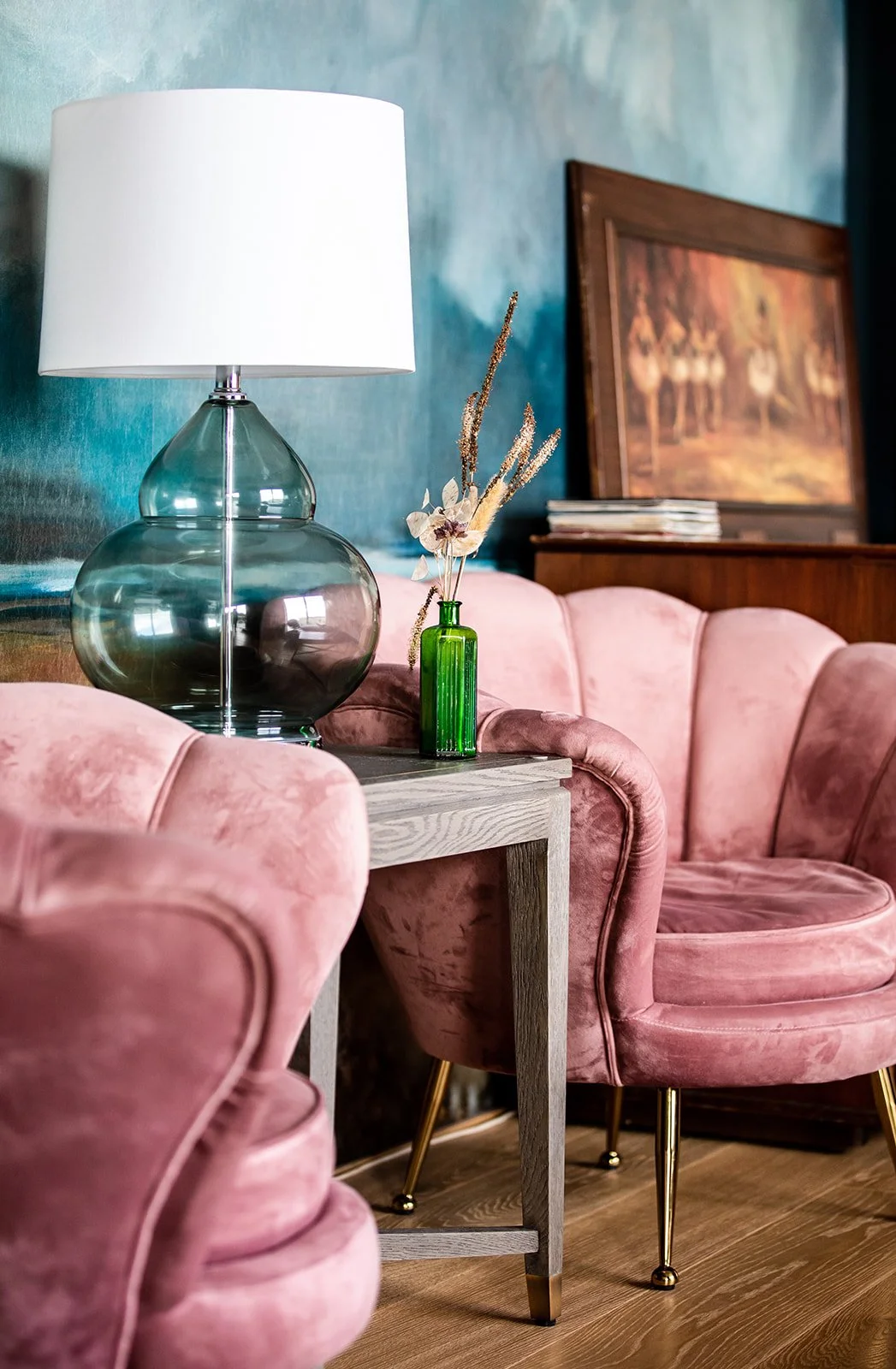 A living room scene with pink velvet armchairs, a side table holding a large glass lamp with a white shade, and a small green vase with dried flowers. In the background is a teal wall with a framed painting and some books.