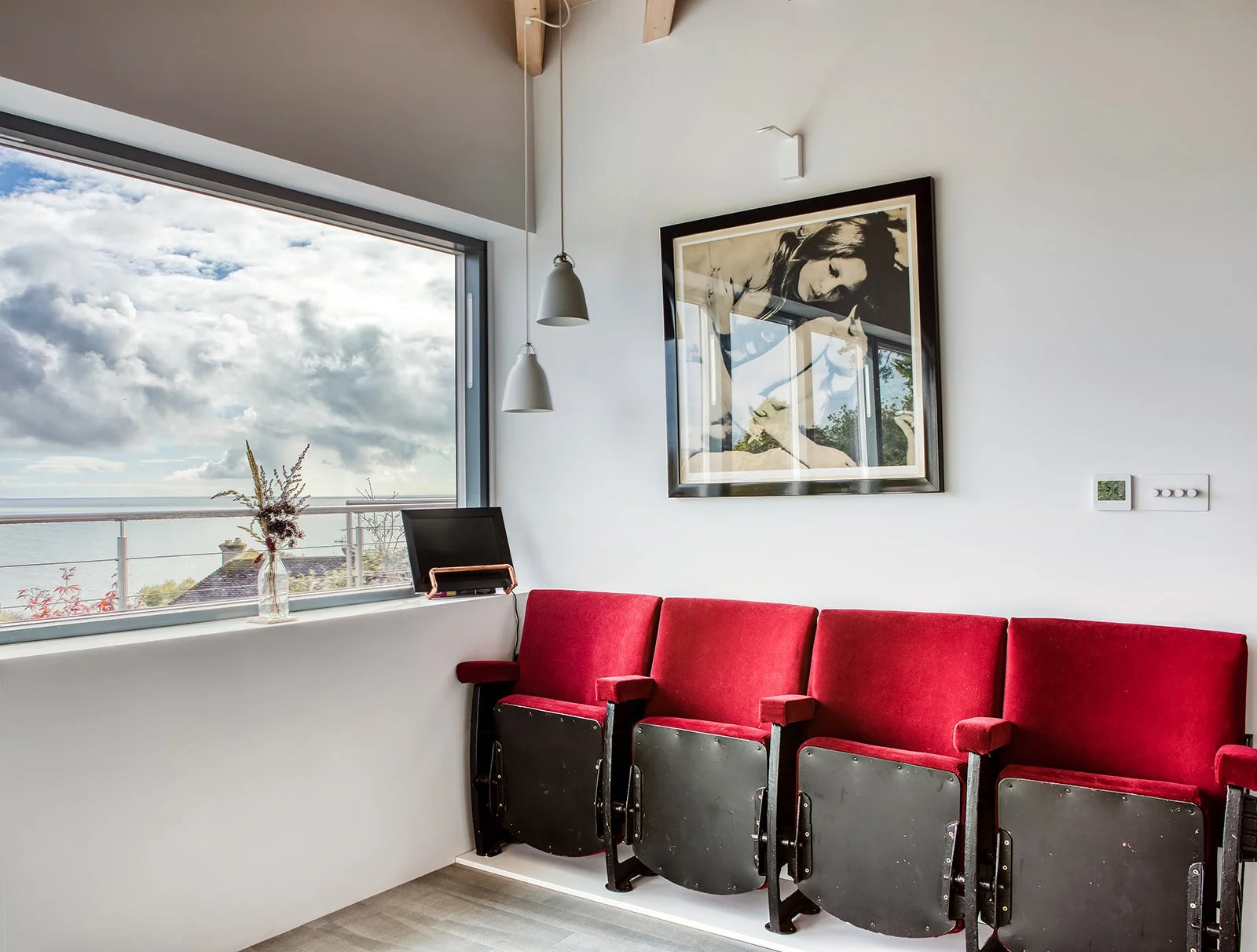 Interior of a room with a large window showing a cloudy sky and ocean view, red theater-style seats lined up against a white wall, a framed black and white photograph, a small monitor on the window ledge, a vase with dried flowers, and two pendant li