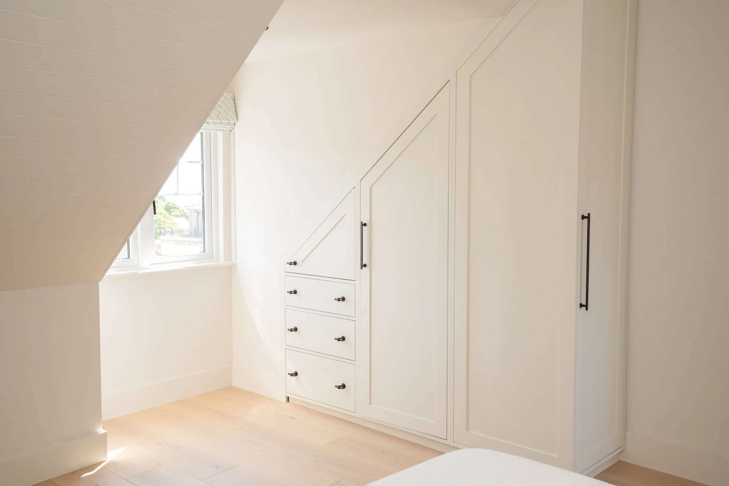 Bright bedroom with a white built-in wardrobe under the sloped ceiling, a window with a view outside, and light wood flooring.
