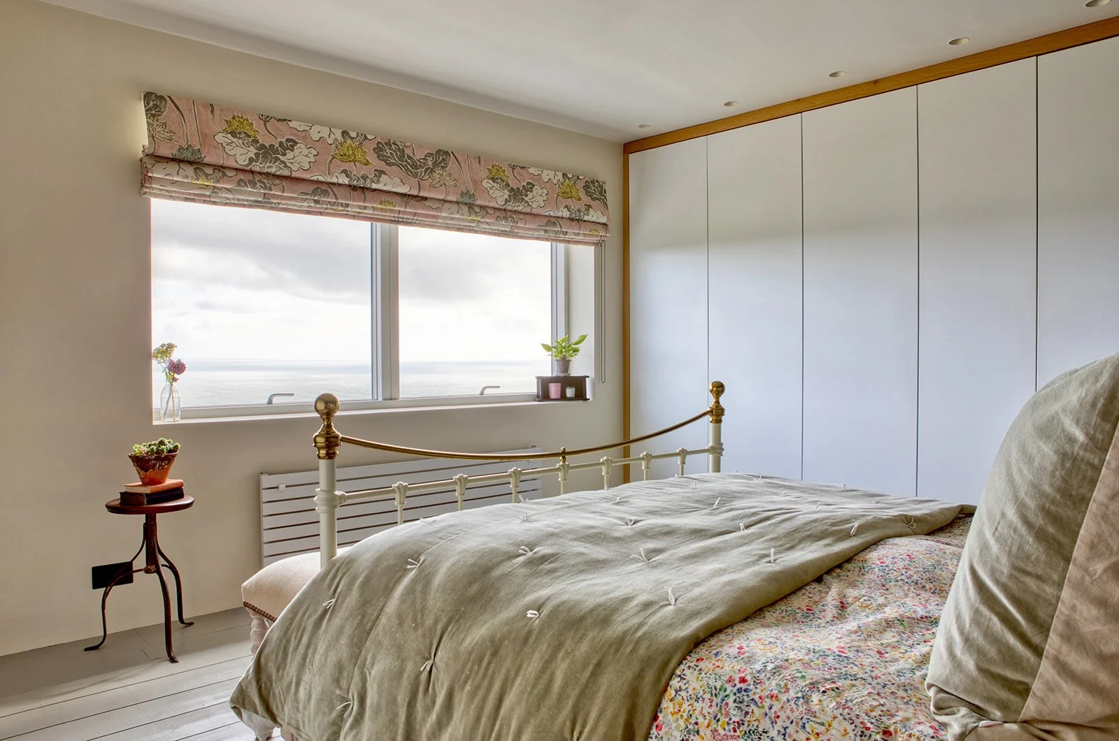 Bedroom with large window overlooking the ocean, beige bed with gray bedding and pillows, small side table with potted cactus, decorative curtains, built-in white wardrobe, and a wooden trim around the ceiling.