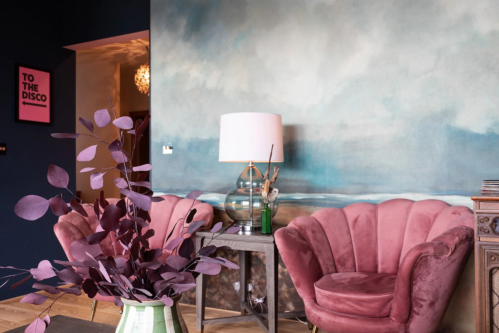 Living room with pink velvet armchairs, a side table with a glass lamp, and a large wall painting of a serene seascape.