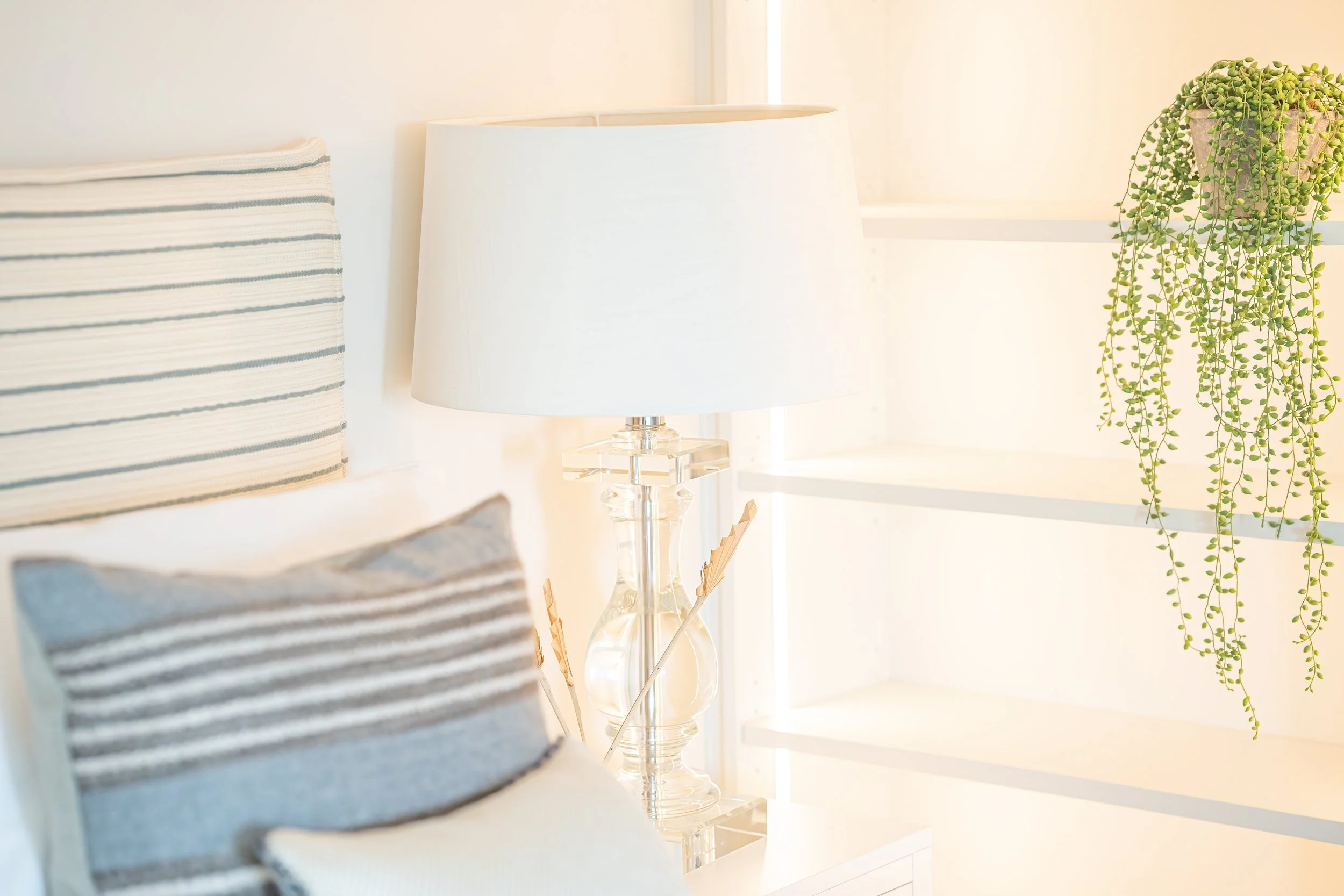 A cozy bedroom corner with a striped pillow, a glass table lamp with a white lampshade, and a hanging green plant in a pot by a window.