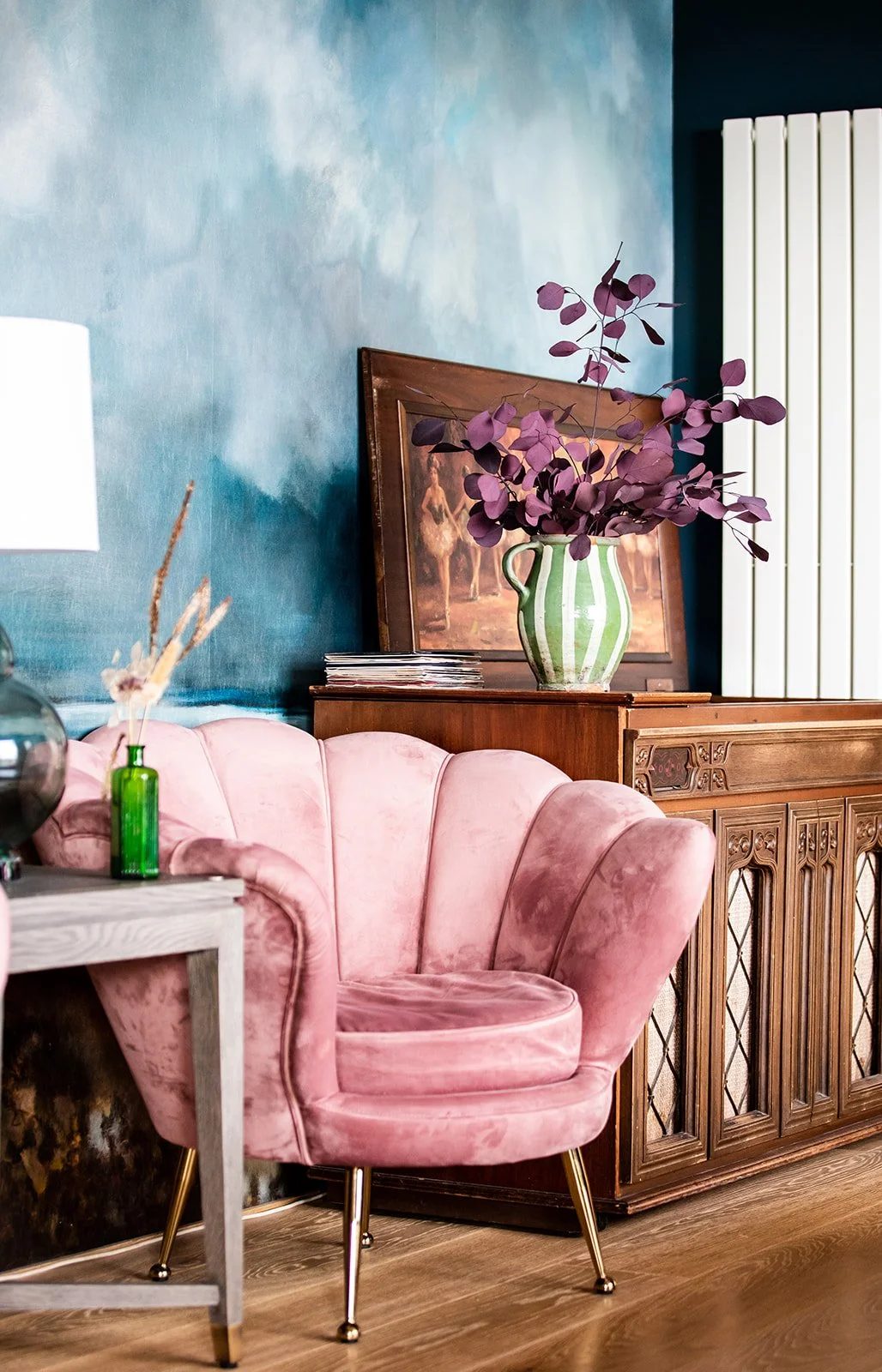 A pink velvet armchair with gold legs next to a wooden sideboard that has a green and white striped vase with purple leaves, a framed artwork, and a stack of magazines, against a blue textured wall.