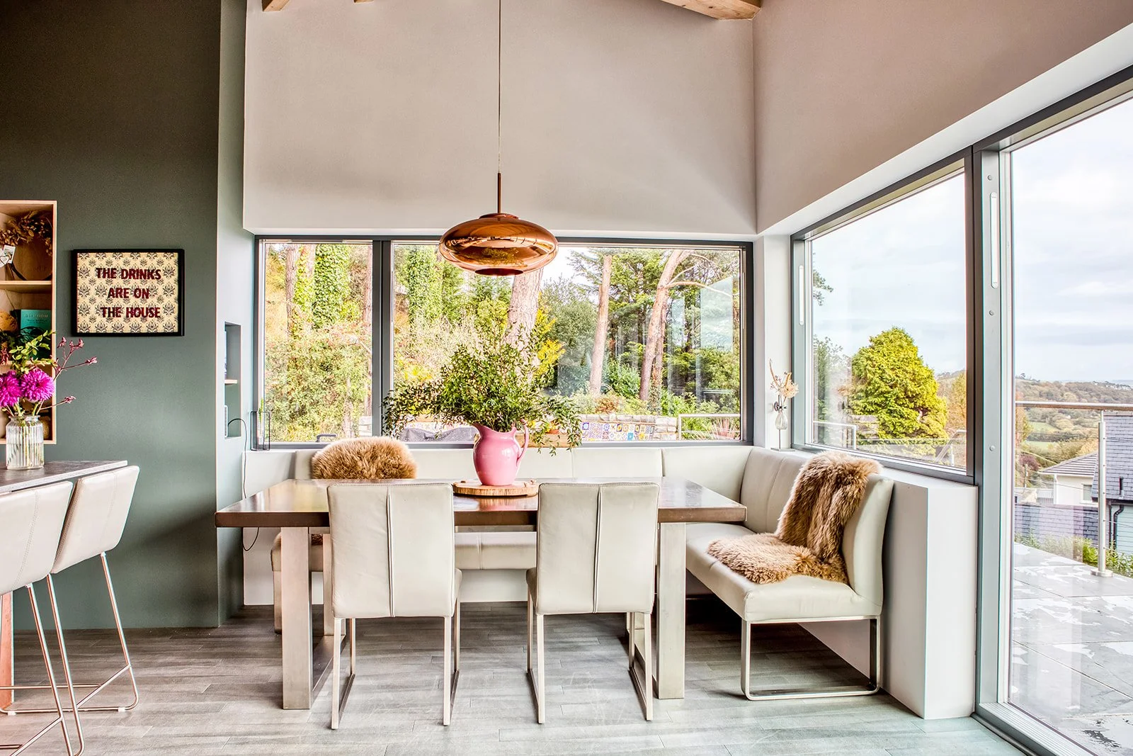 Bright dining area with a wooden table, surrounded by cream-colored chairs, a pink vase with green foliage, and a large window offering a scenic view of trees and the outdoors.
