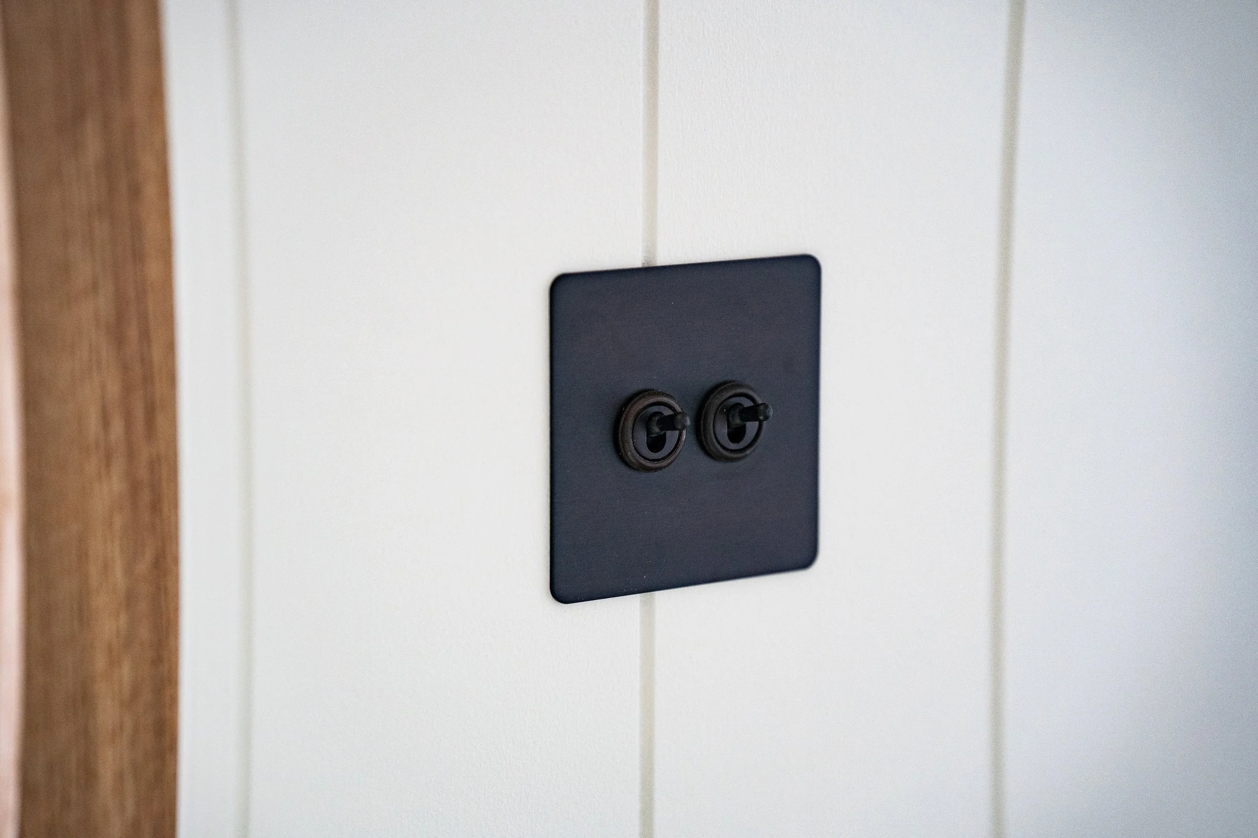 Close-up of a black wall plate with two toggle switches on a white paneled wall, with a wooden strip on the left.