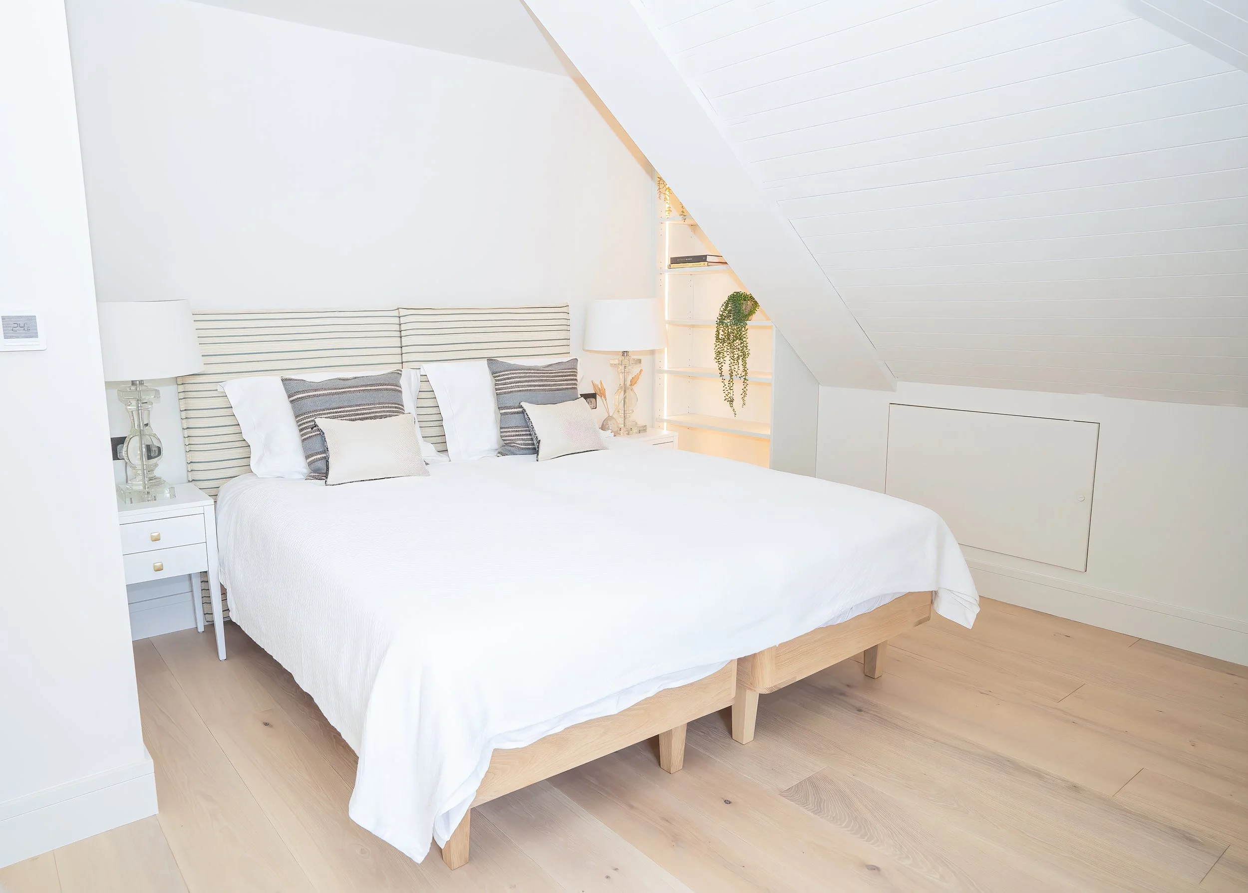 A cozy bedroom with a white bed, striped headboard, and several pillows. There are bedside lamps, a built-in bookshelf with plants and books, and light wood flooring.