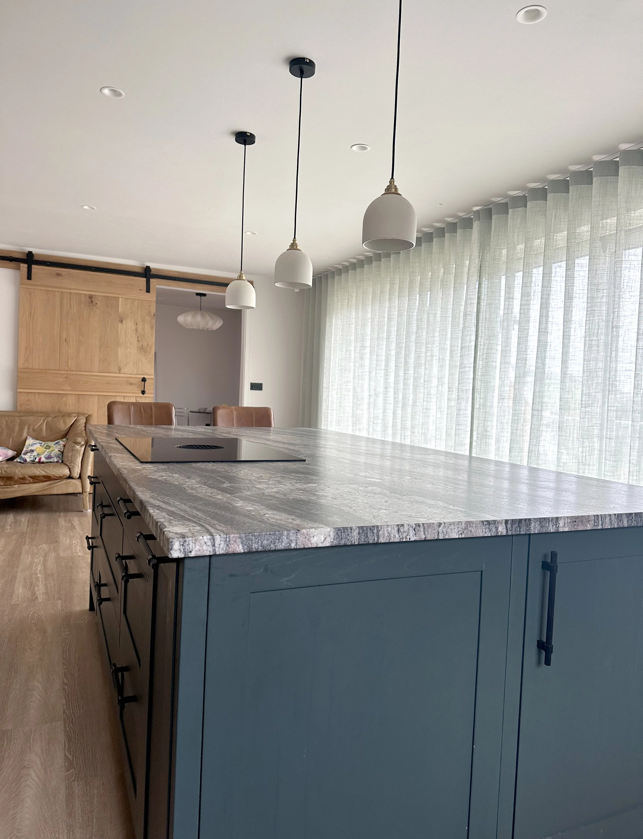 Modern kitchen with a large marble-topped island, pendant lights, wooden cabinetry, and sheer curtains covering large windows.