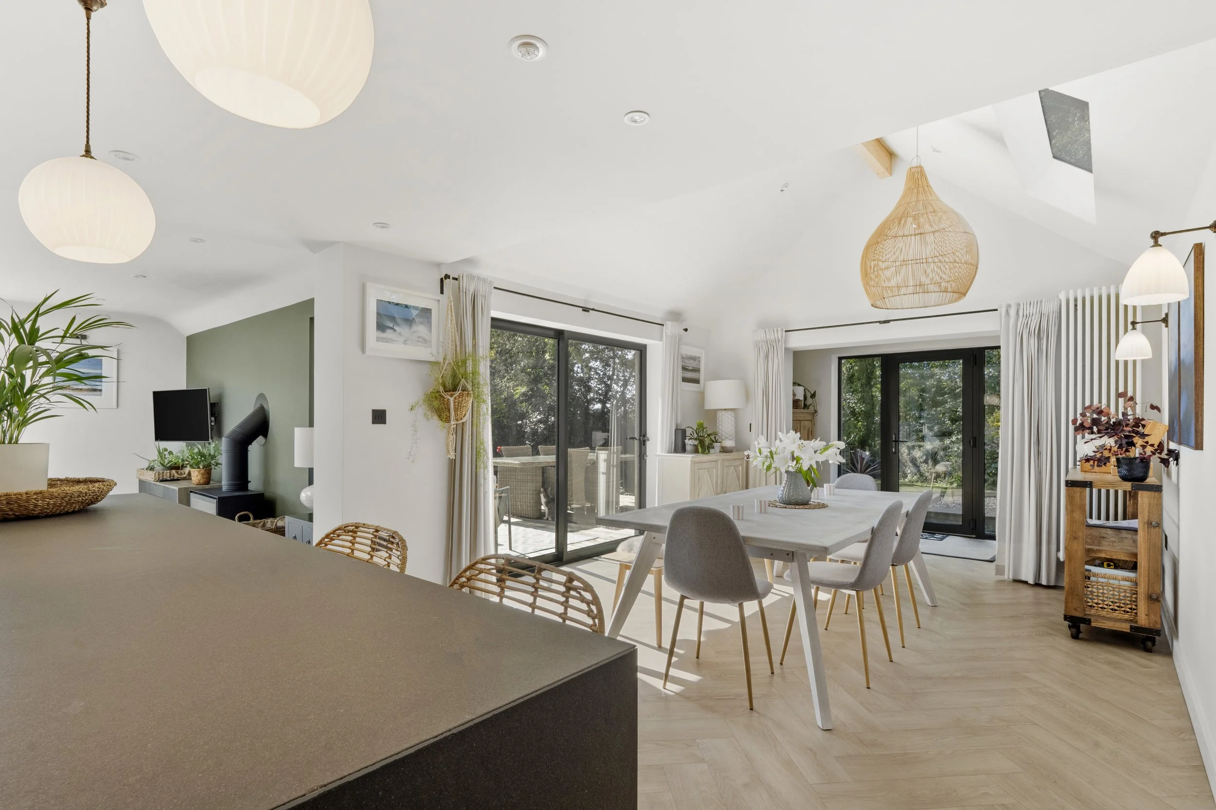 Open-concept living and dining area with white walls, light wooden floors, a white dining table, gray chairs, and large sliding glass doors leading to an outdoor patio. Decor includes hanging wicker light fixtures, potted plants, artwork, and a sideb