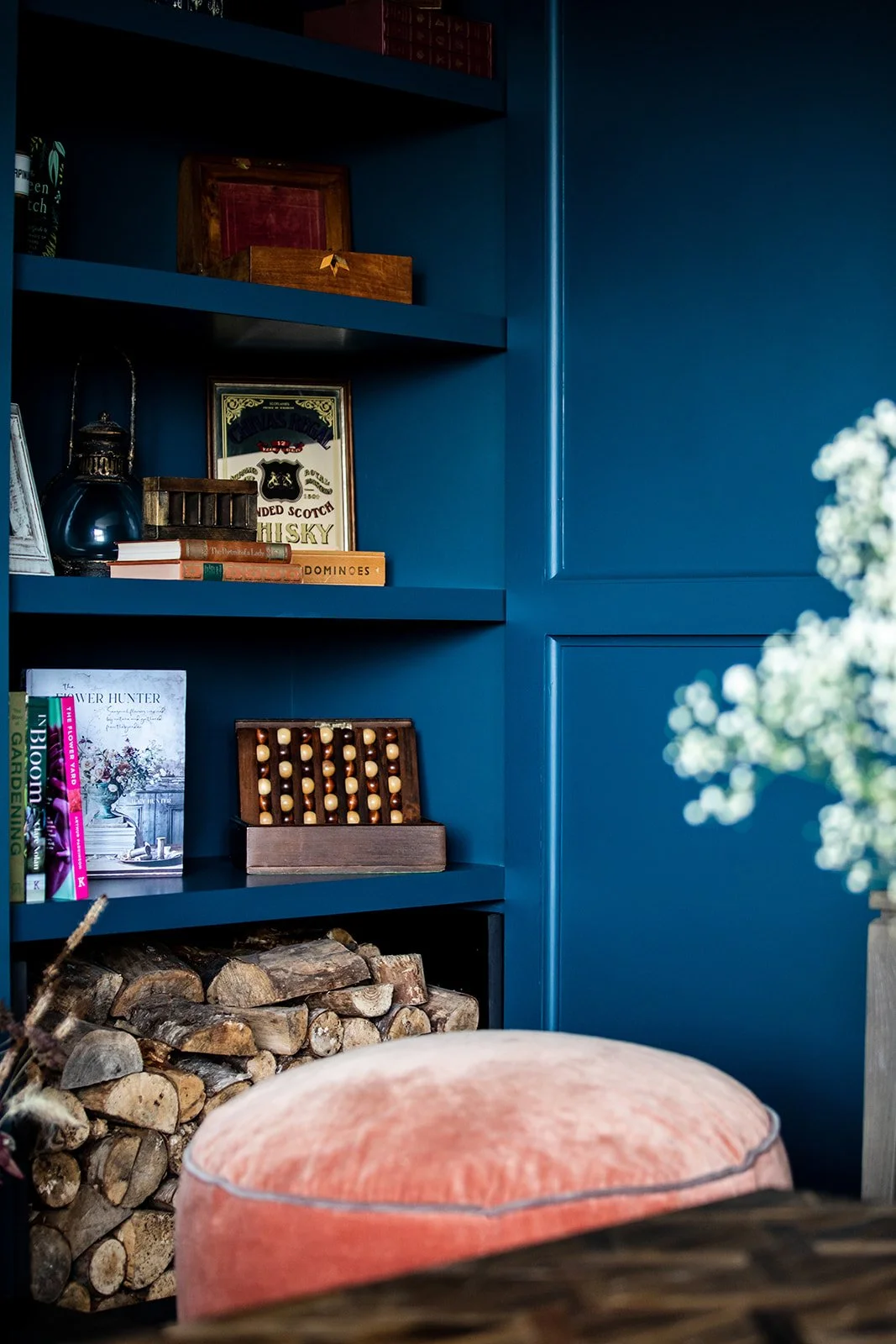 A cozy corner of a room with a dark blue built-in bookshelf filled with books and decorative items, a crammed wood storage beneath, a pink velvet ottoman, and a tall vase with white flowers on the right.