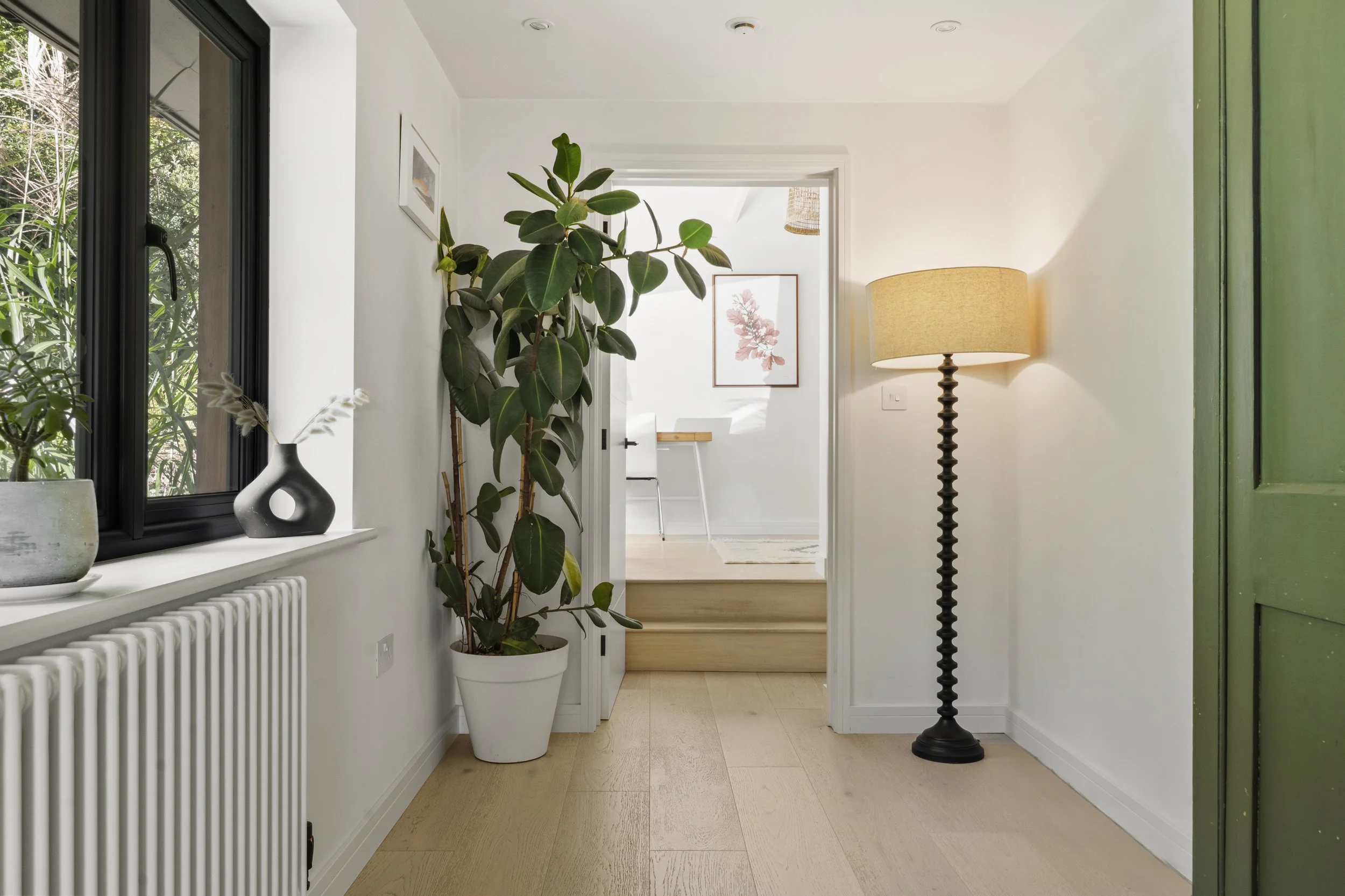 A cozy interior space with a large window, potted plant, decorative vases, a floor lamp, and a view into a bright room with artwork and a desk.