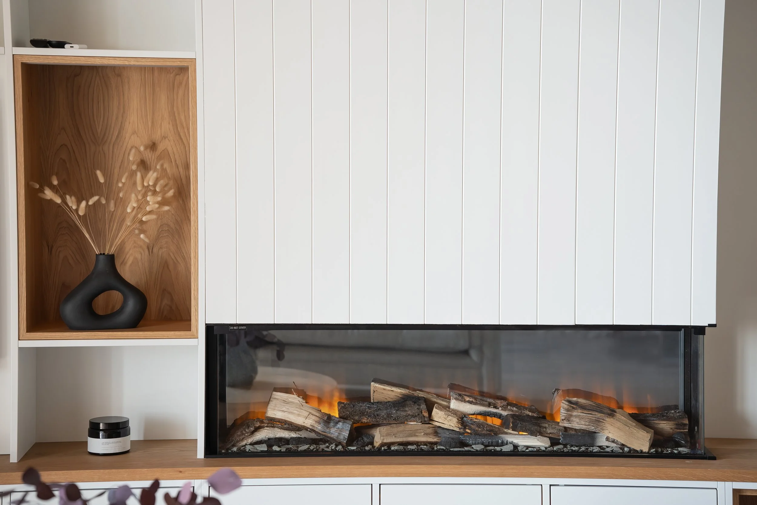 Modern electric fireplace with realistic logs, set into a wooden and white wall unit, with decorative black vase and a small jar nearby.