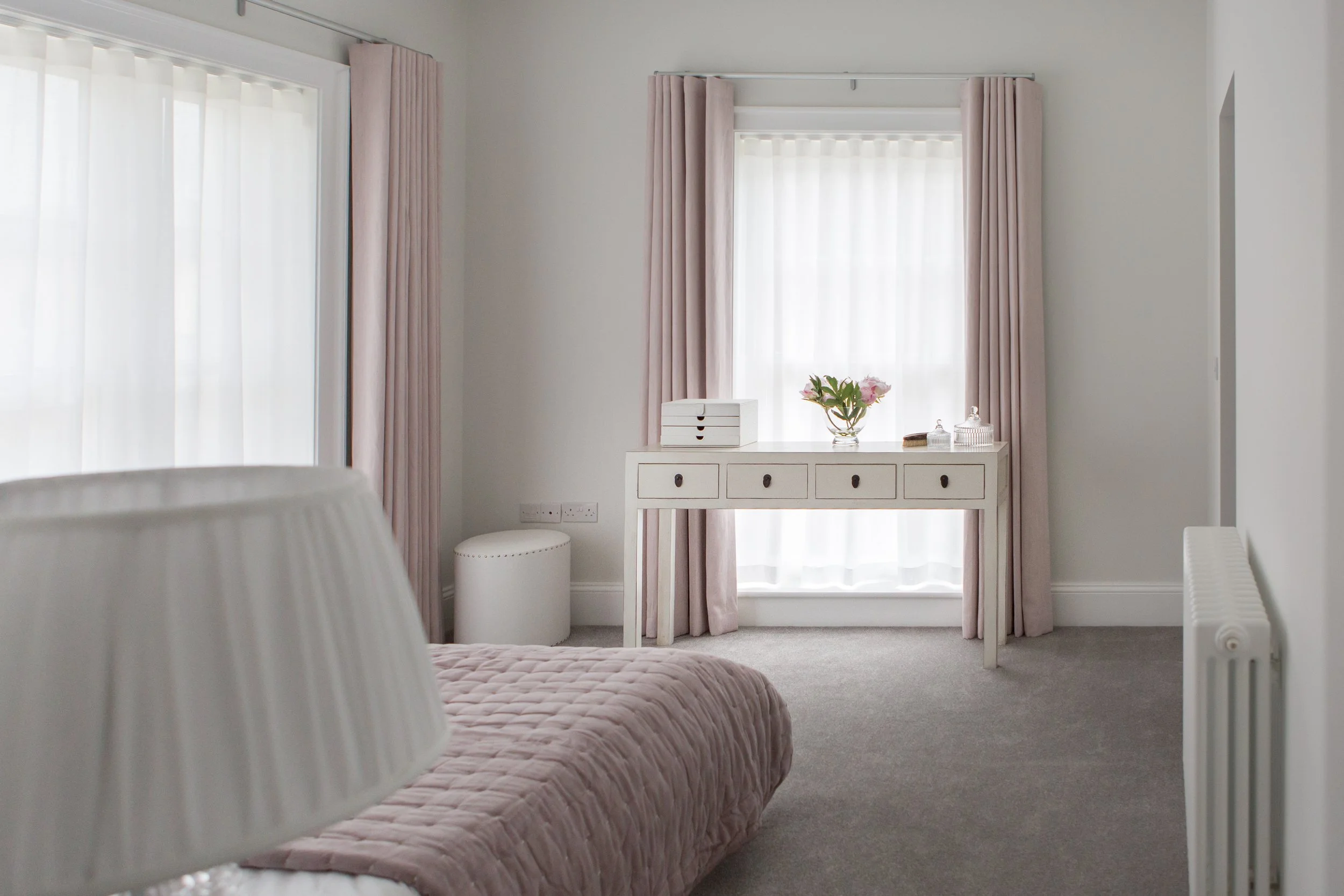 A bedroom with a bed covered in a pink quilt, white walls, a white dresser with drawers, a vase of flowers, and pink curtains on two windows.