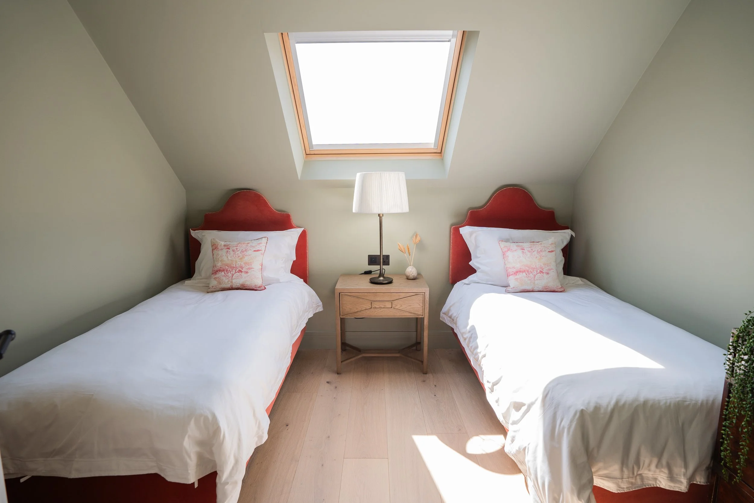 Bedroom with two single beds with red headboards, pillows, and white bedding, a wooden nightstand with a lamp and decorative items, and a skylight window on the sloped ceiling letting in sunlight.