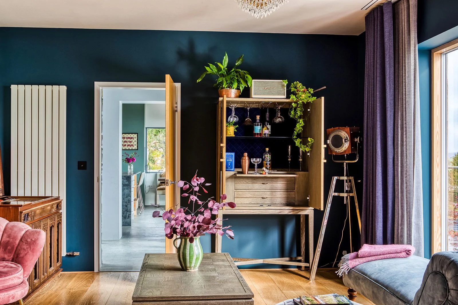Living room with dark blue wall, wooden furniture, a pink velvet armchair, a coffee table with a purple flower arrangement, and a large window with purple curtains.