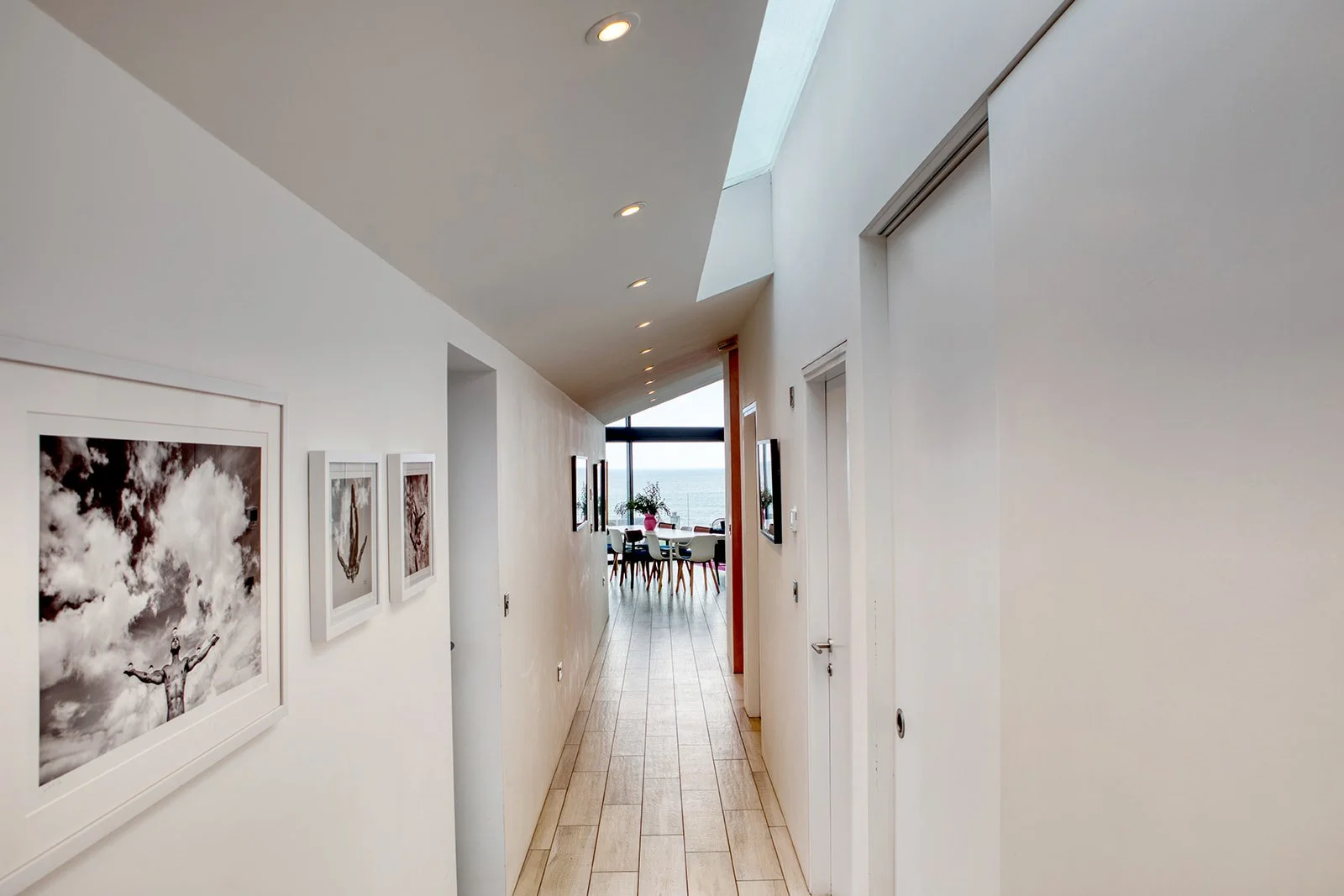 Bright hallway with white walls, wooden floor, and hanging framed black-and-white photographs, leading to a dining area with a view of the ocean outside.