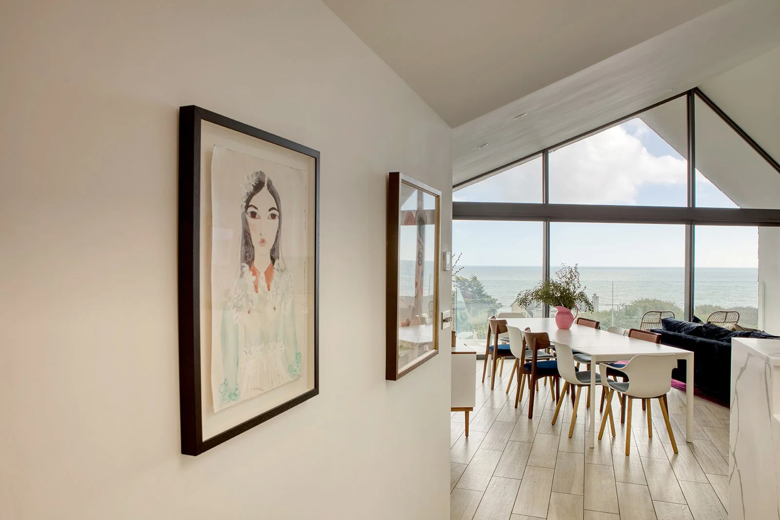 Room with large window overlooking the ocean, with dining table and chairs, and artwork of a woman on the wall.